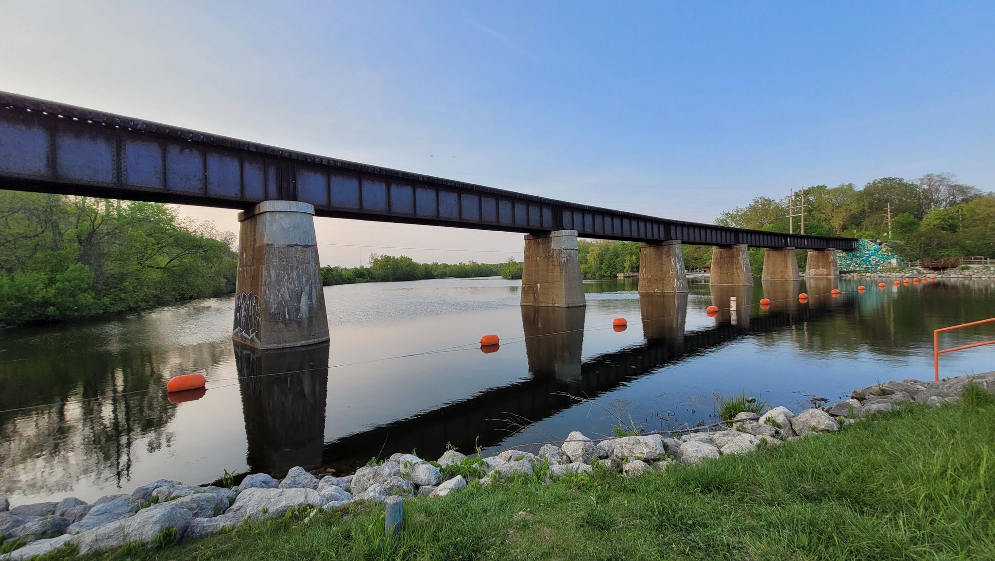 A long metal train bridge spans a wide river under a clear blue sky. - Parker Mill County Park – Argo Dam Railway Bridge loop from Arbor Hills Nature Area