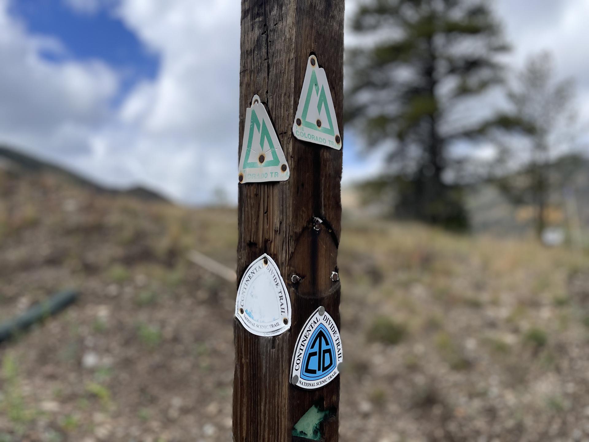 Several trail markers are affixed to a weathered wooden post outdoors.