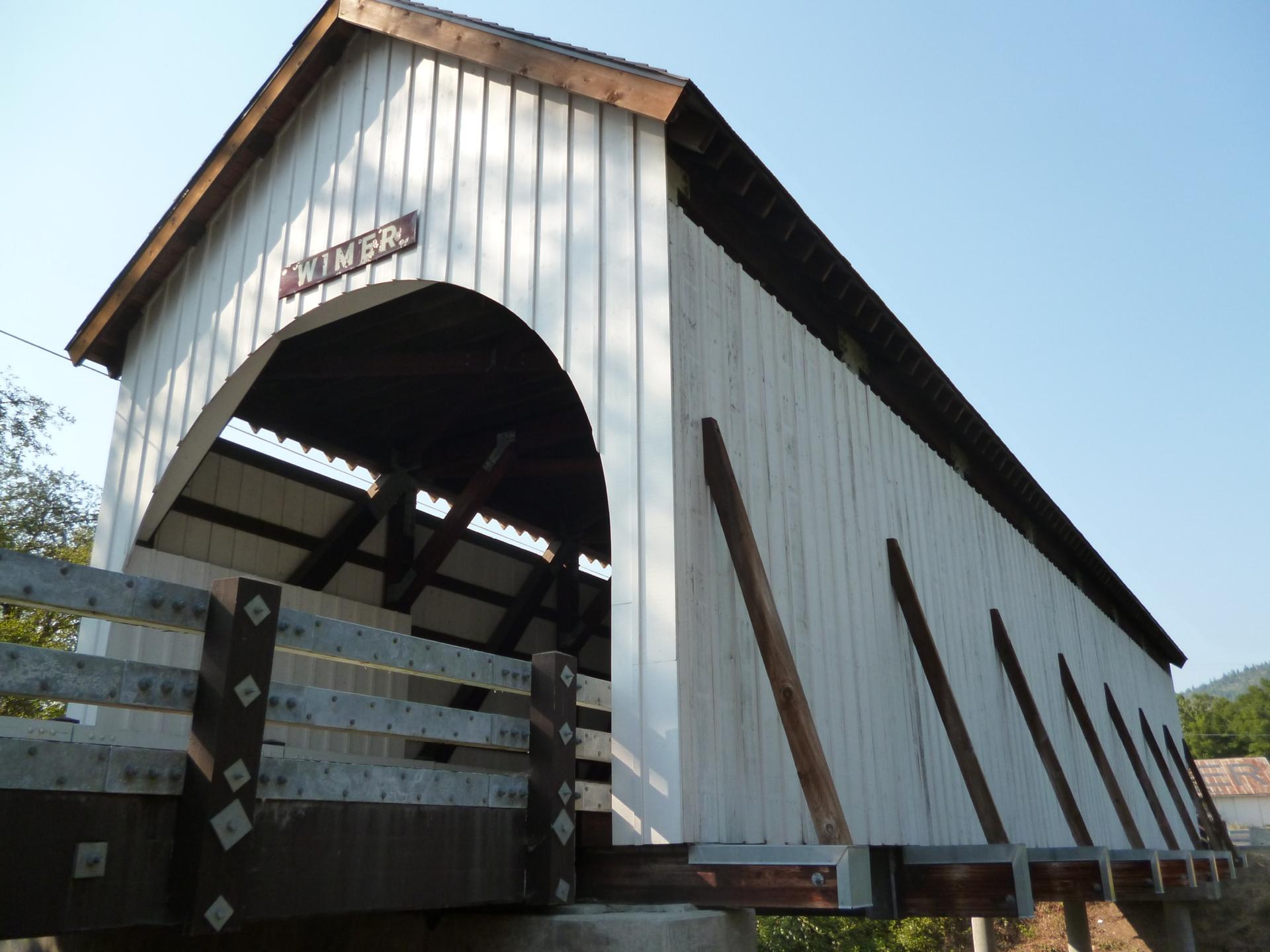 A white wooden covered bridge with an arched entrance under bright sunlight.