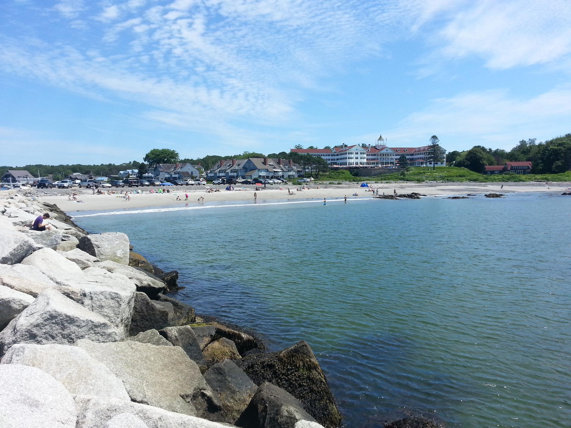 Sunny beach with people, blue water, and distant buildings from rocky shore.