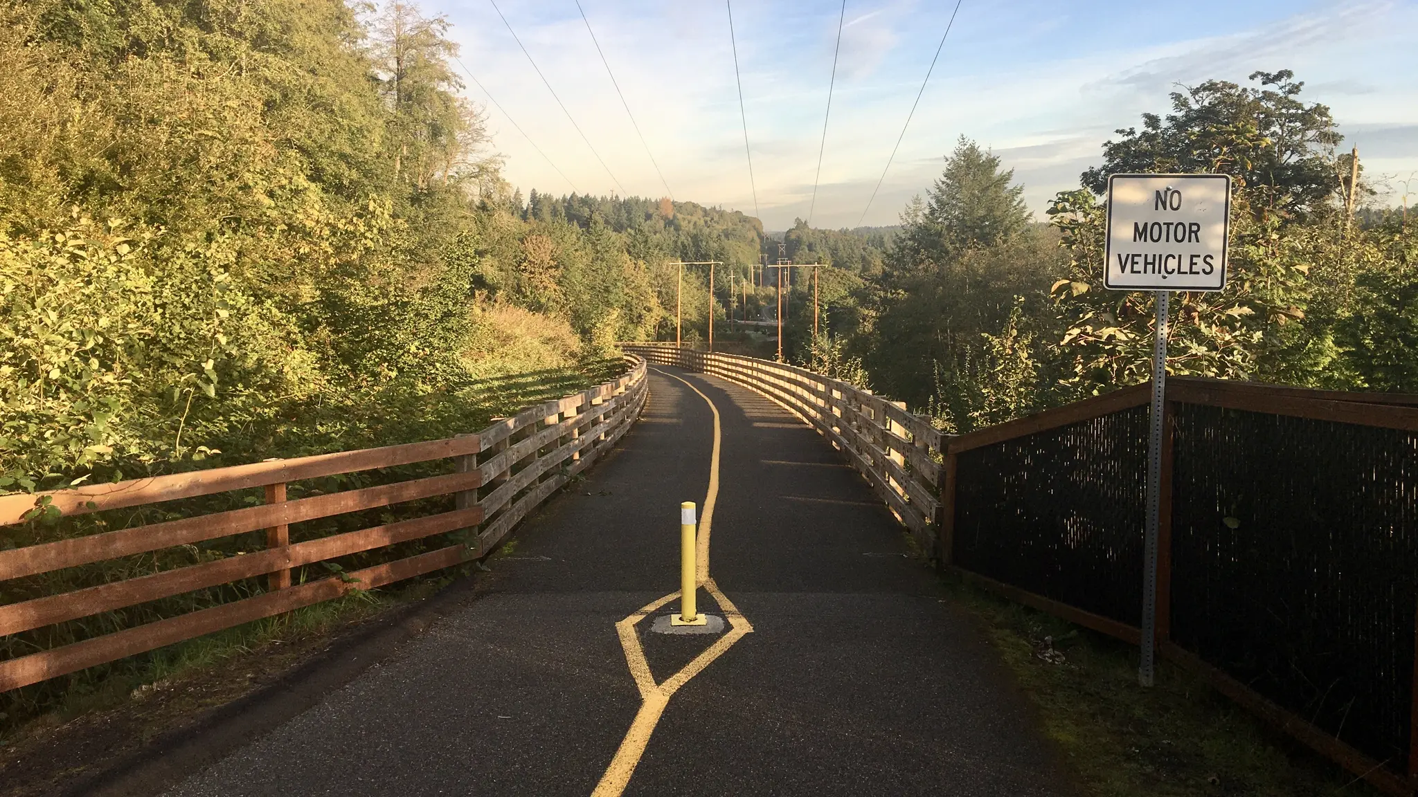 Paved path through green woods with fences and a "No Motor Vehicles" sign.