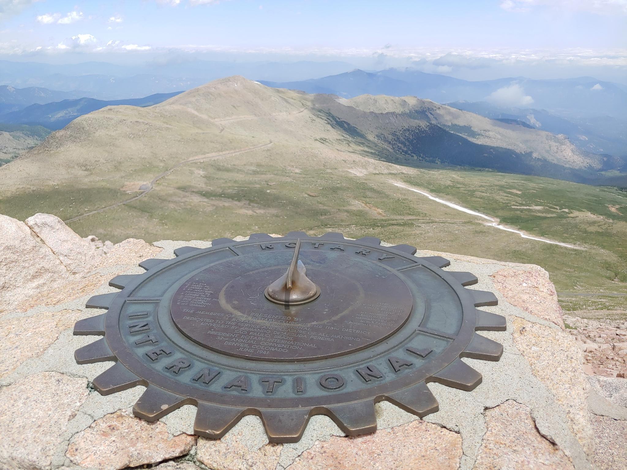 Metal sundial on rocks overlooking vast, arid mountains under a clear sky.