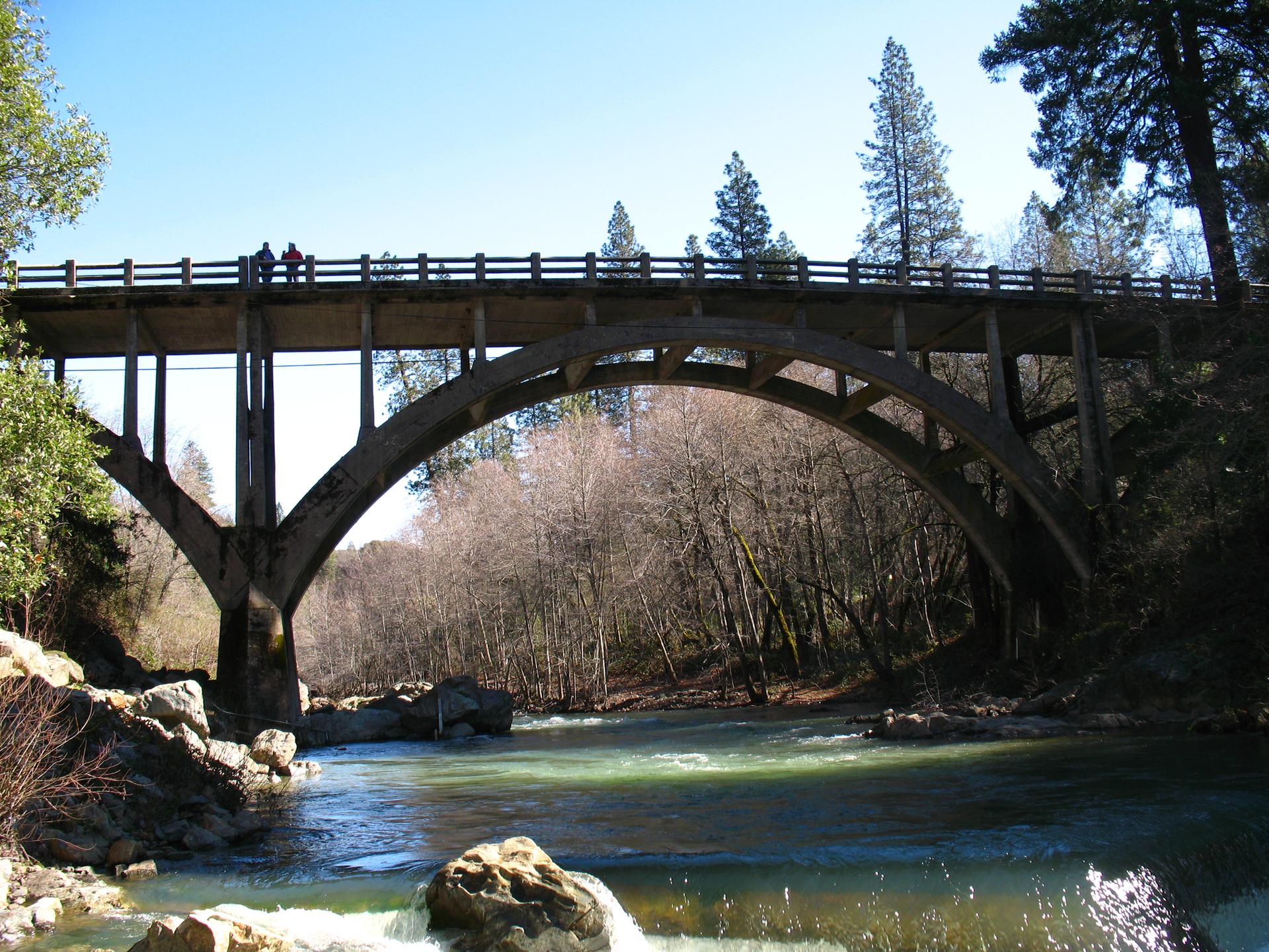 A large arched bridge spans a river in a sunny, forested valley.