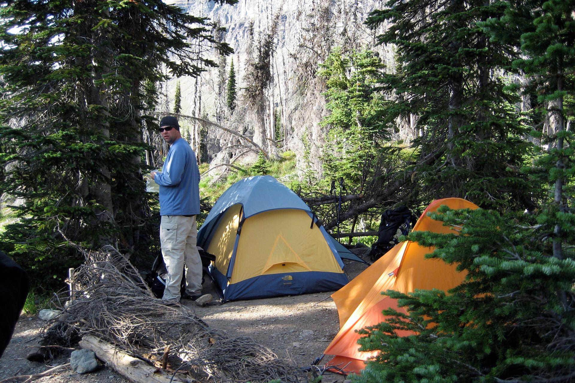 Man standing near two colorful tents in a sunlit forest campsite.