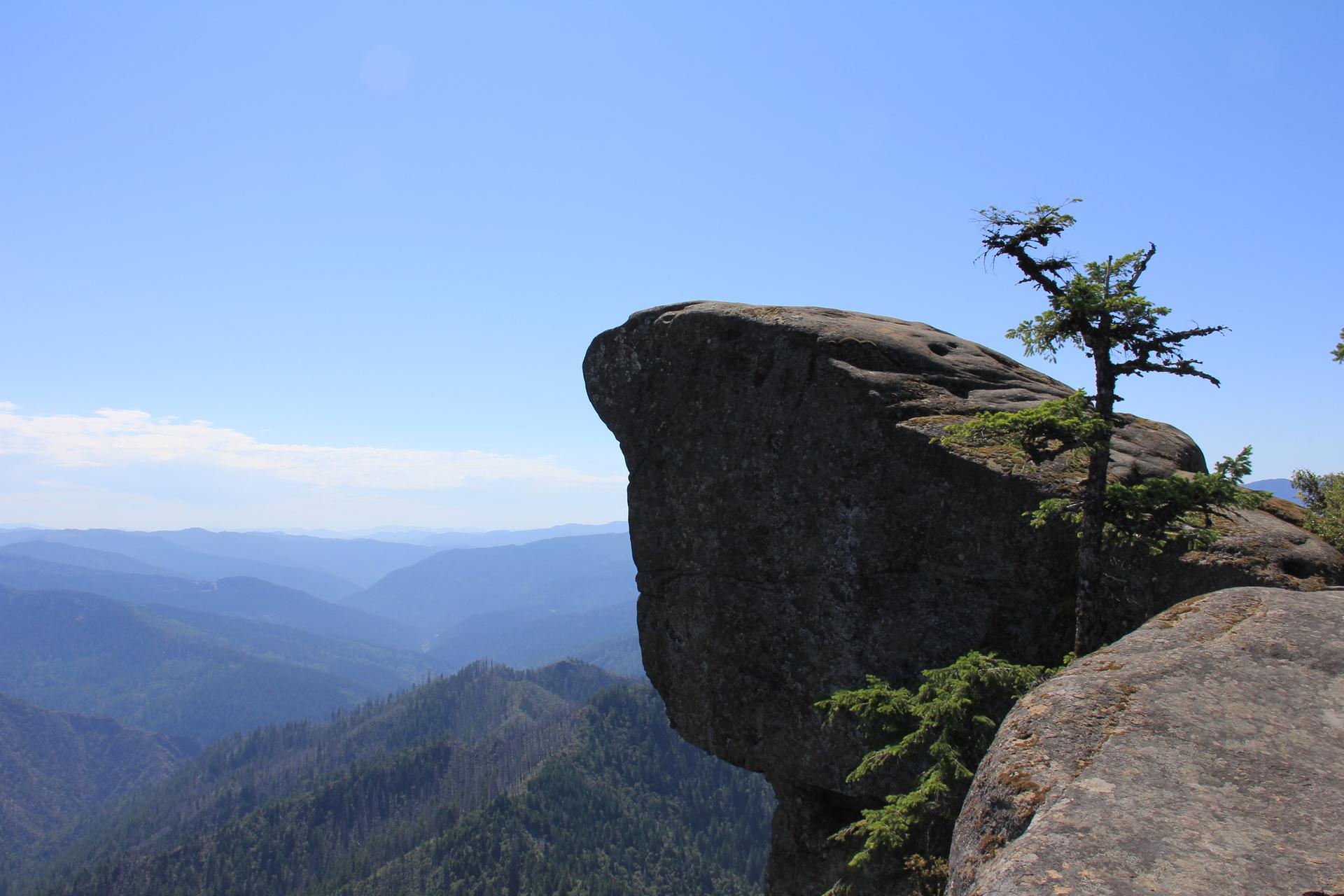 A large rock overhang with a small tree overlooks distant forested mountains under a clear sky.