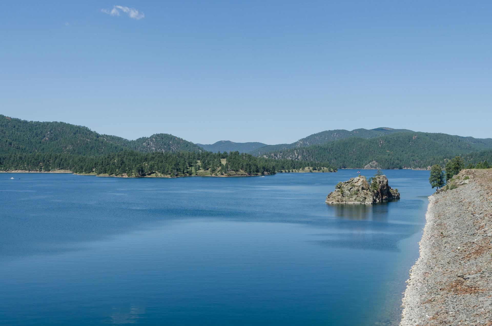 Sunny day over a vast blue lake, rocky island, and distant green mountains.