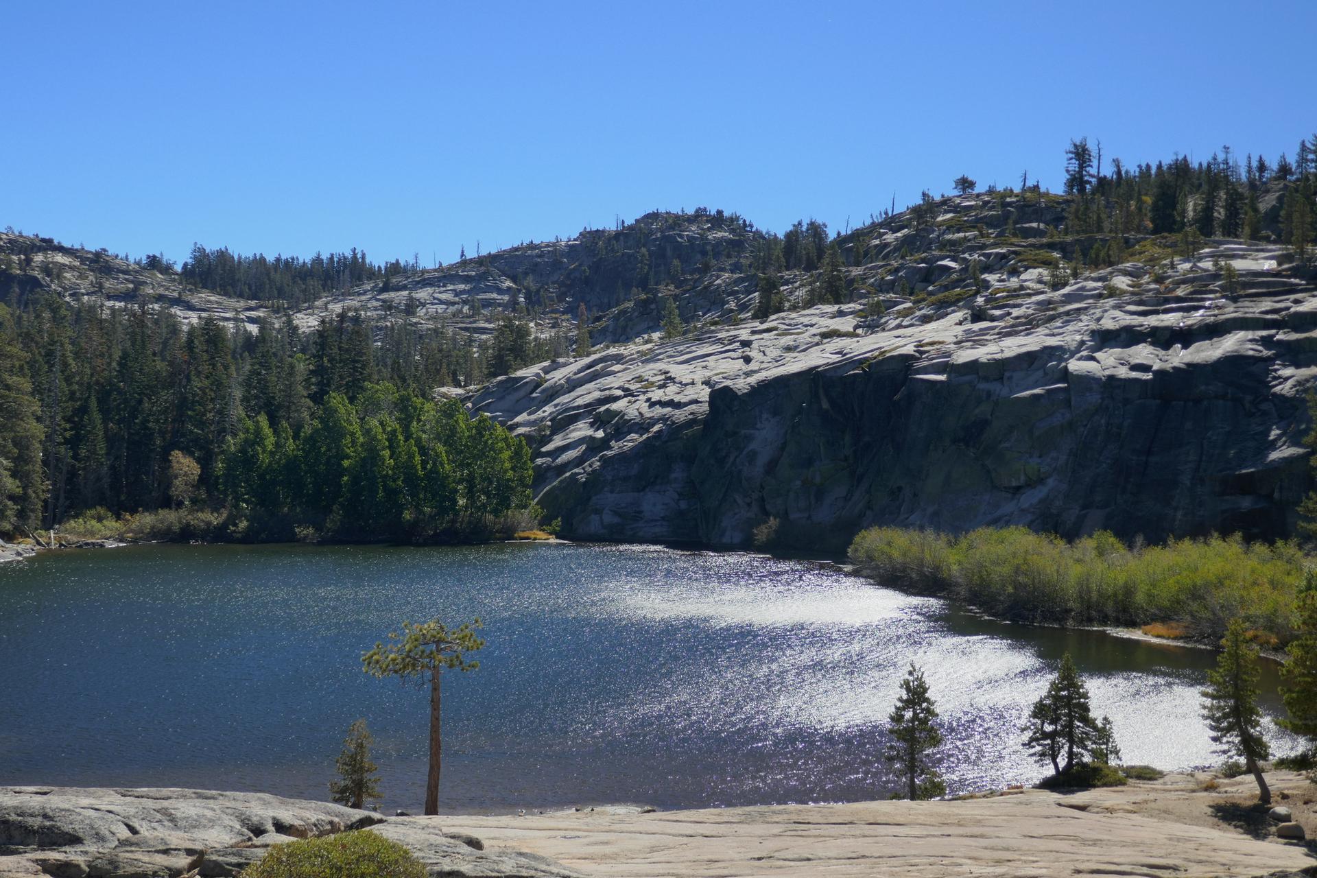 Bright lake with sun glinting, surrounded by rocky mountains and green trees.