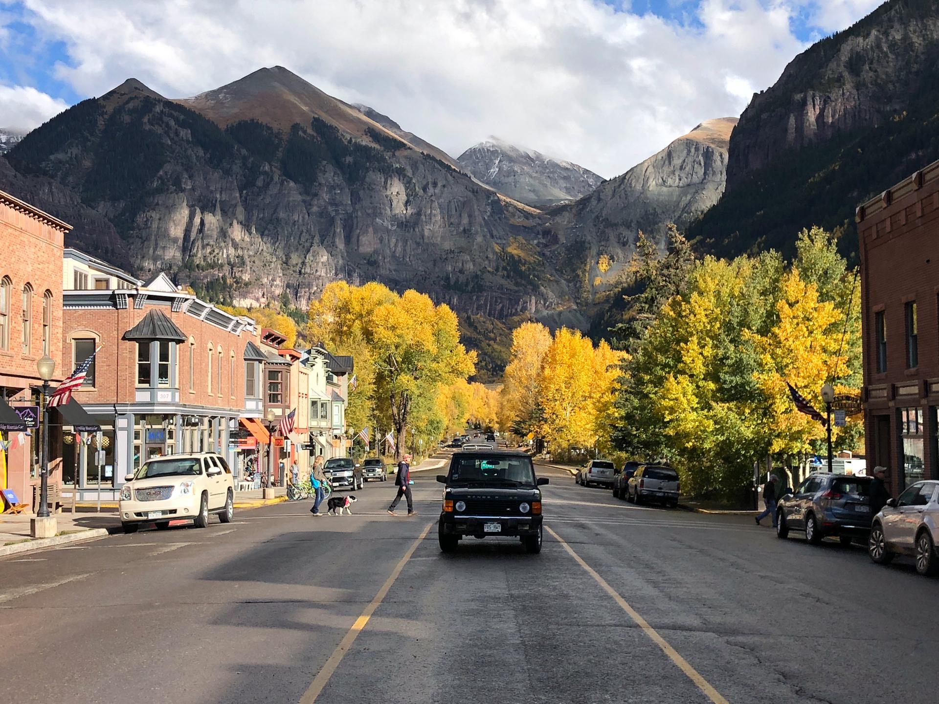Street with autumn trees, buildings, cars, and towering mountains under a bright sky.