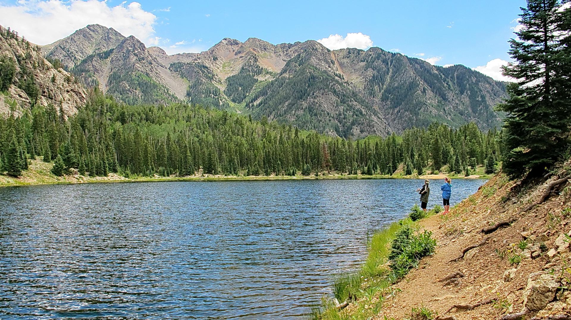 Two people stand by a clear mountain lake surrounded by forests and peaks on a bright day.