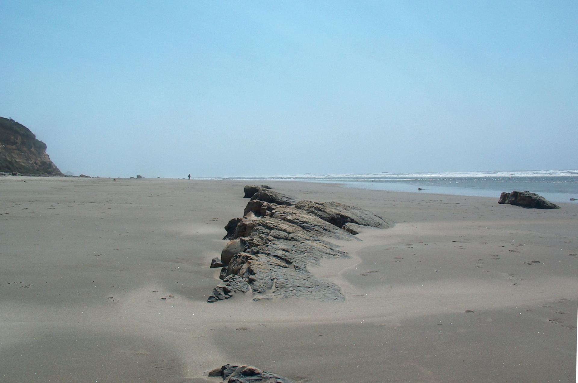 Large rocks on a vast sandy beach with ocean waves and a distant person.
