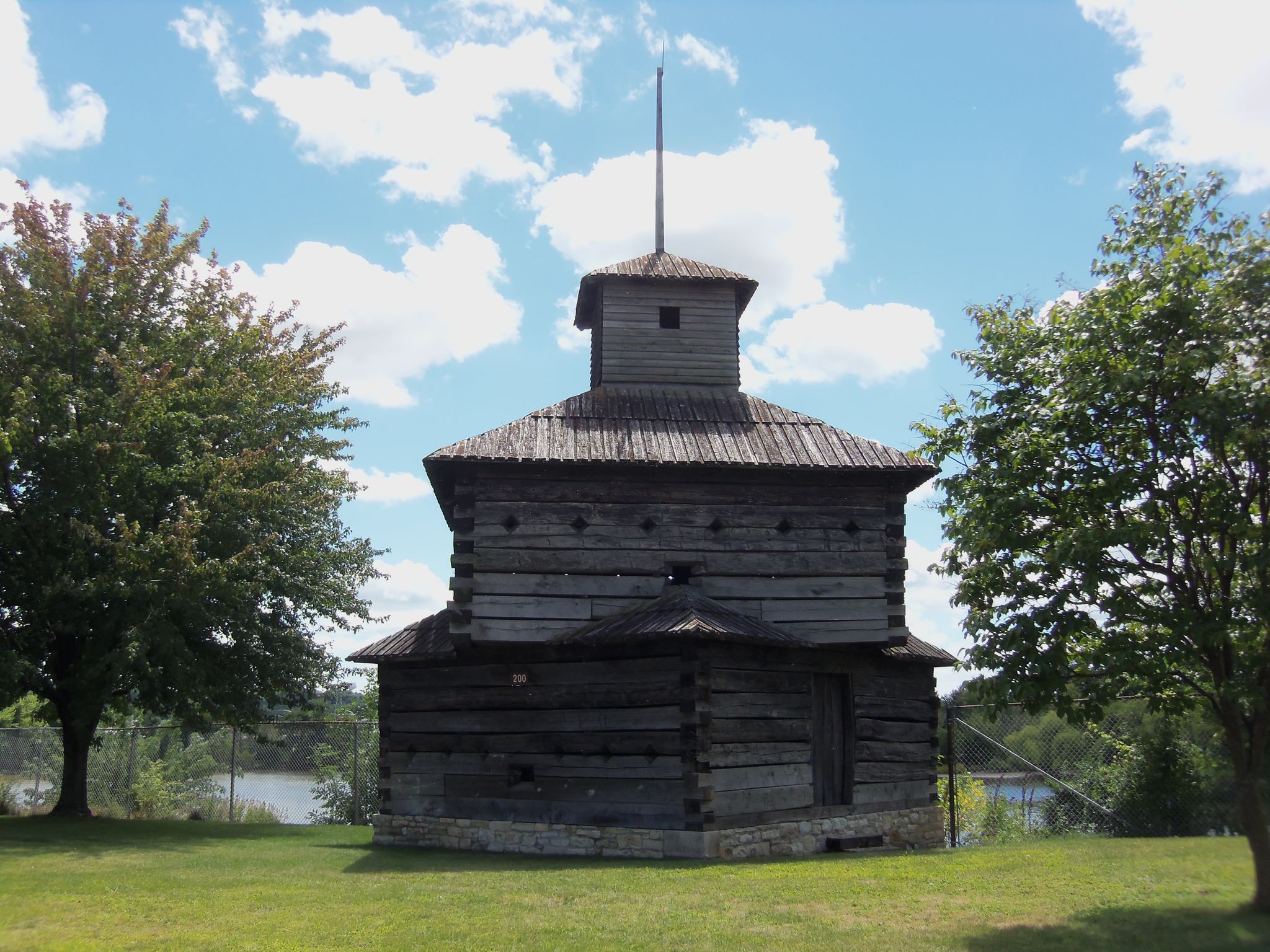 Rustic wooden blockhouse on a grassy hill overlooking water under a sunny sky. - Riverfront Trail to Fort Armstrong