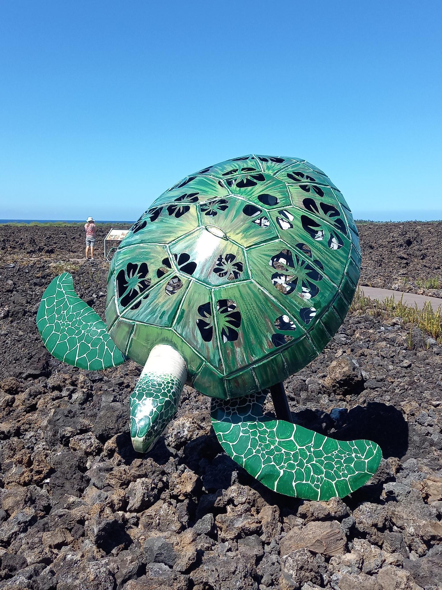 Large green turtle sculpture on dark rocks, with a person in the distance.