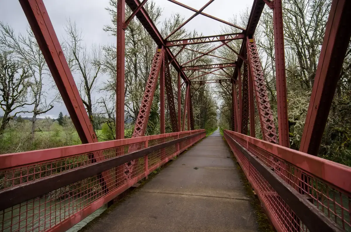 Red metal truss bridge with a path, bare trees, under an overcast sky.