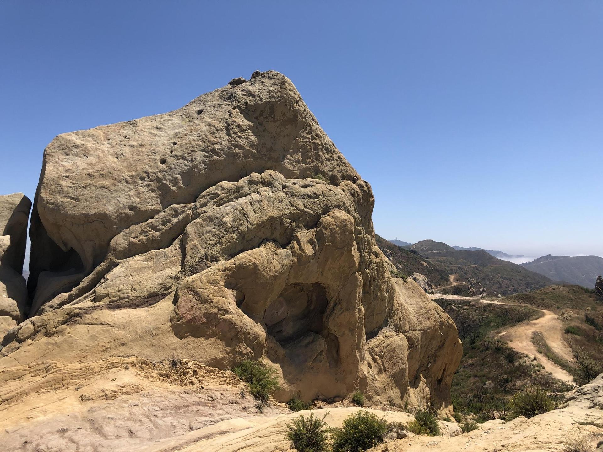 Large, weathered rock formation overlooking a mountainous landscape under blue sky.