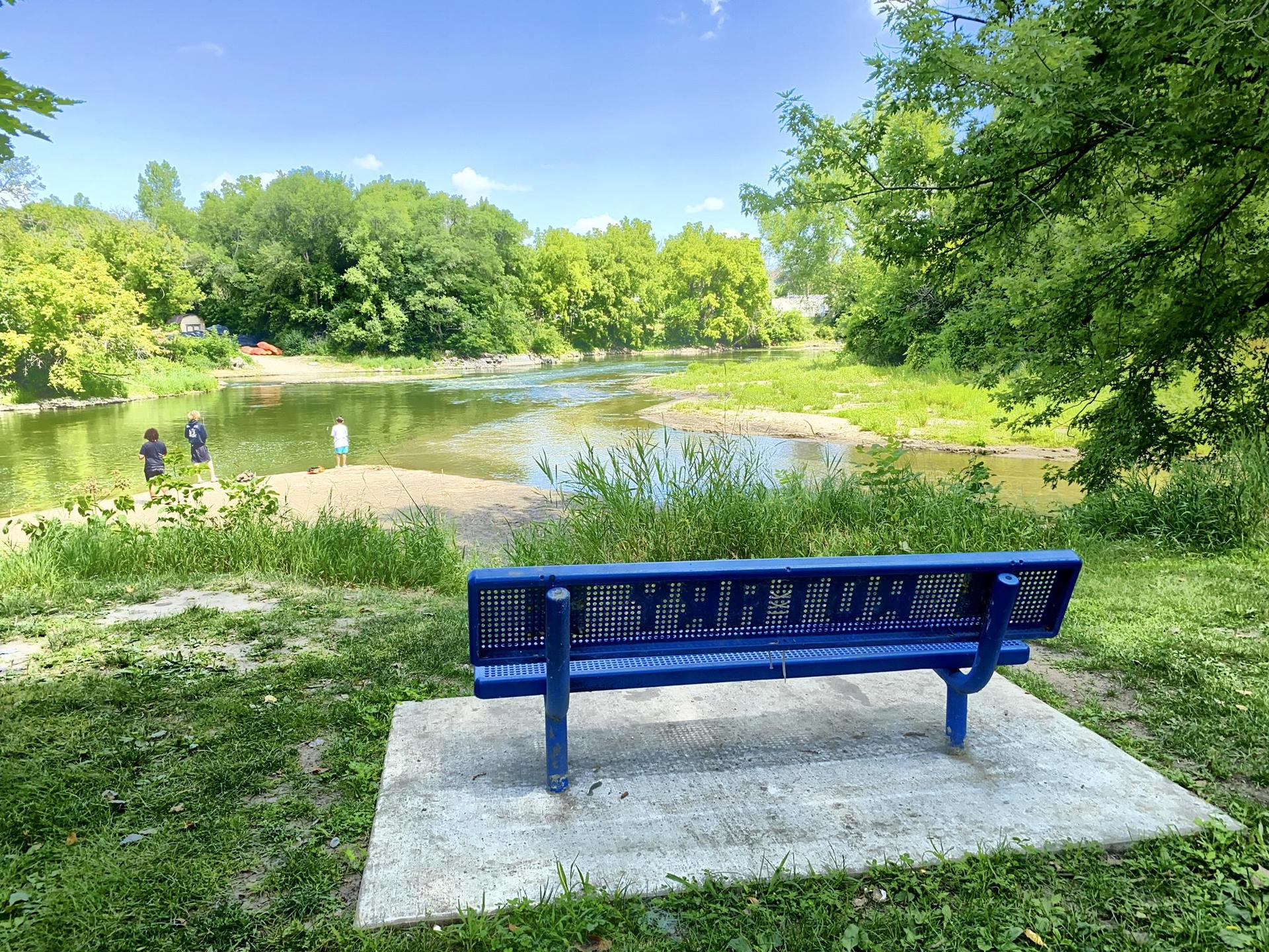 Blue park bench overlooking a sunny river with people on the sandy bank.
