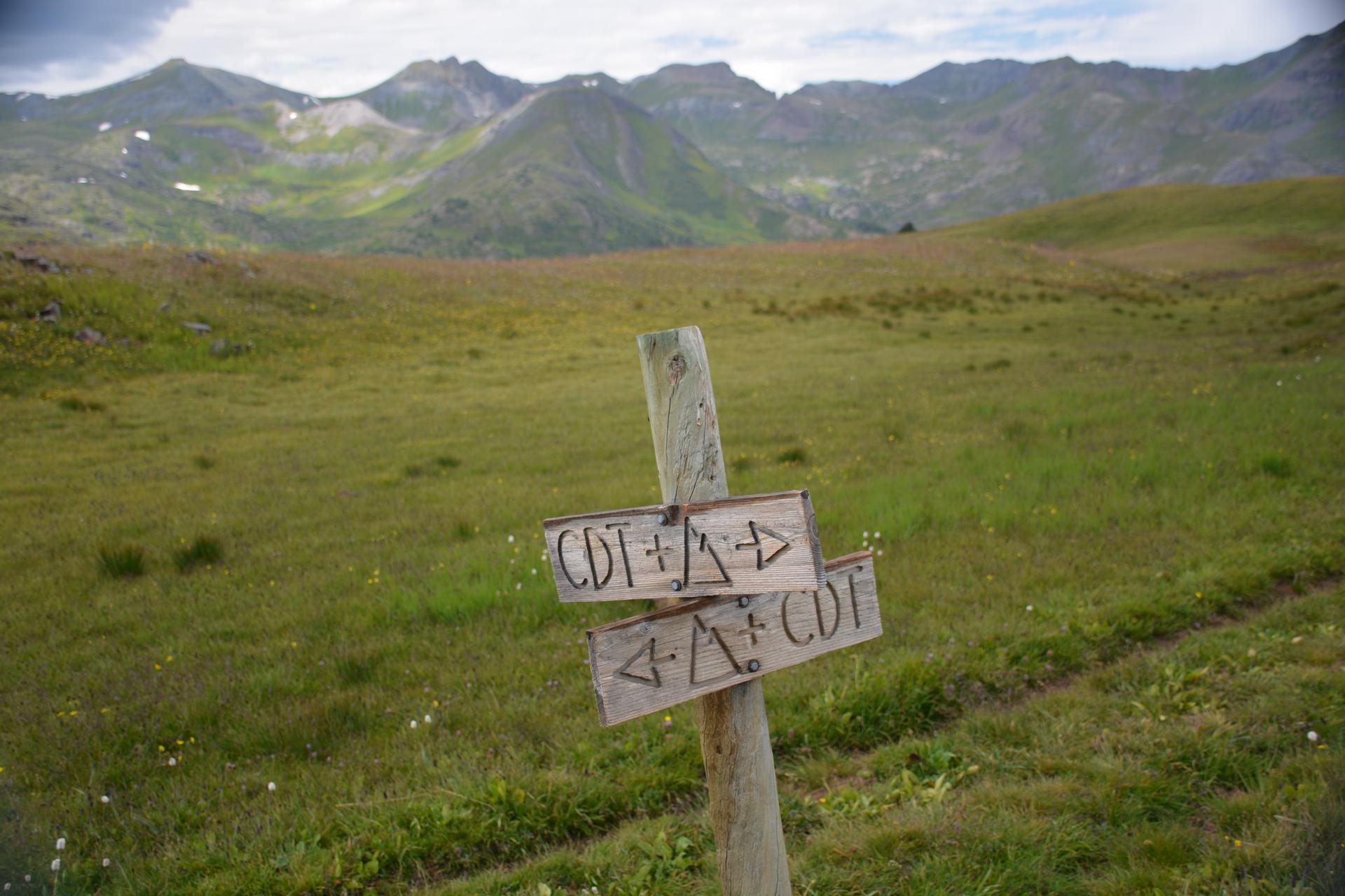 Wooden trail sign in a green mountain meadow under a cloudy sky.