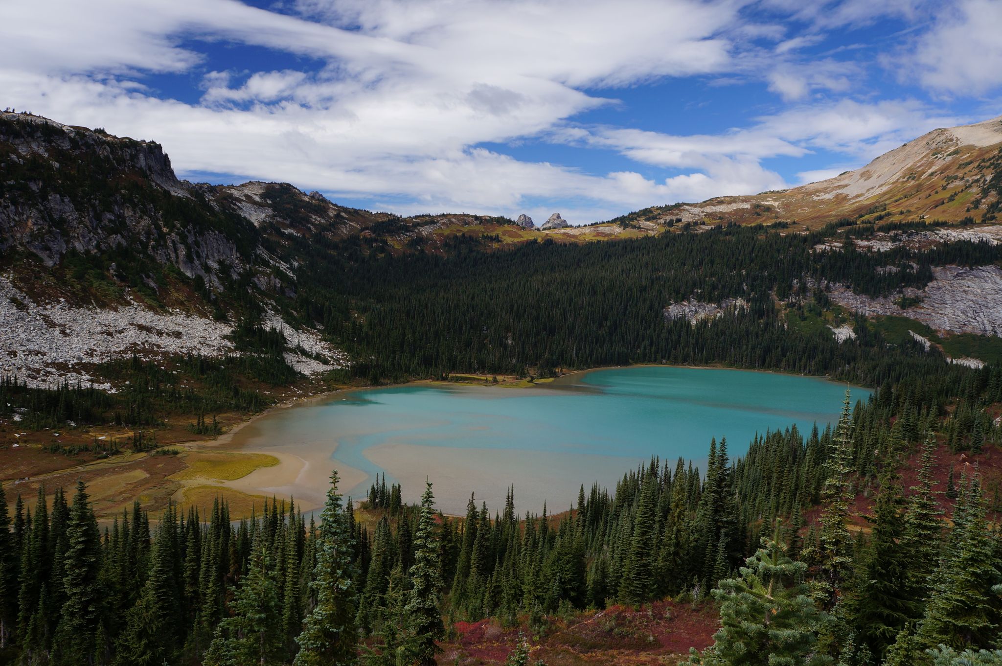 A vibrant turquoise lake nestled among evergreen forests and rocky mountains under clouds. - Cloudy Pass Trail