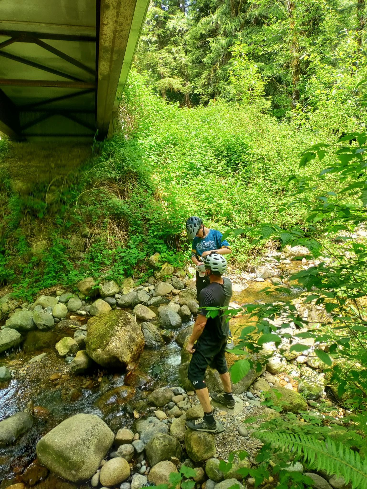 Two people in helmets stand in a rocky stream under a bridge.