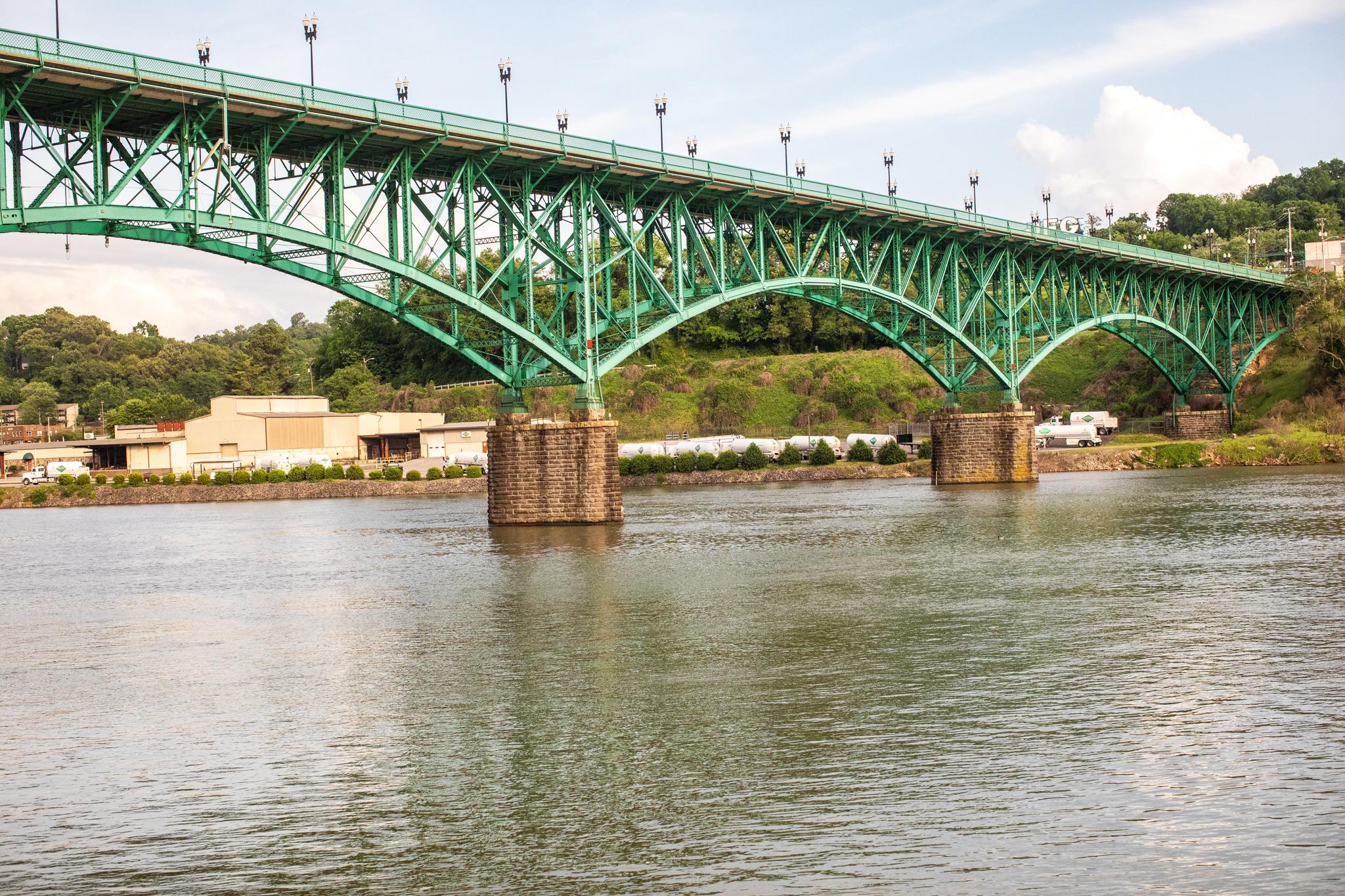 A large green metal arch bridge spans a wide river under a bright sky.