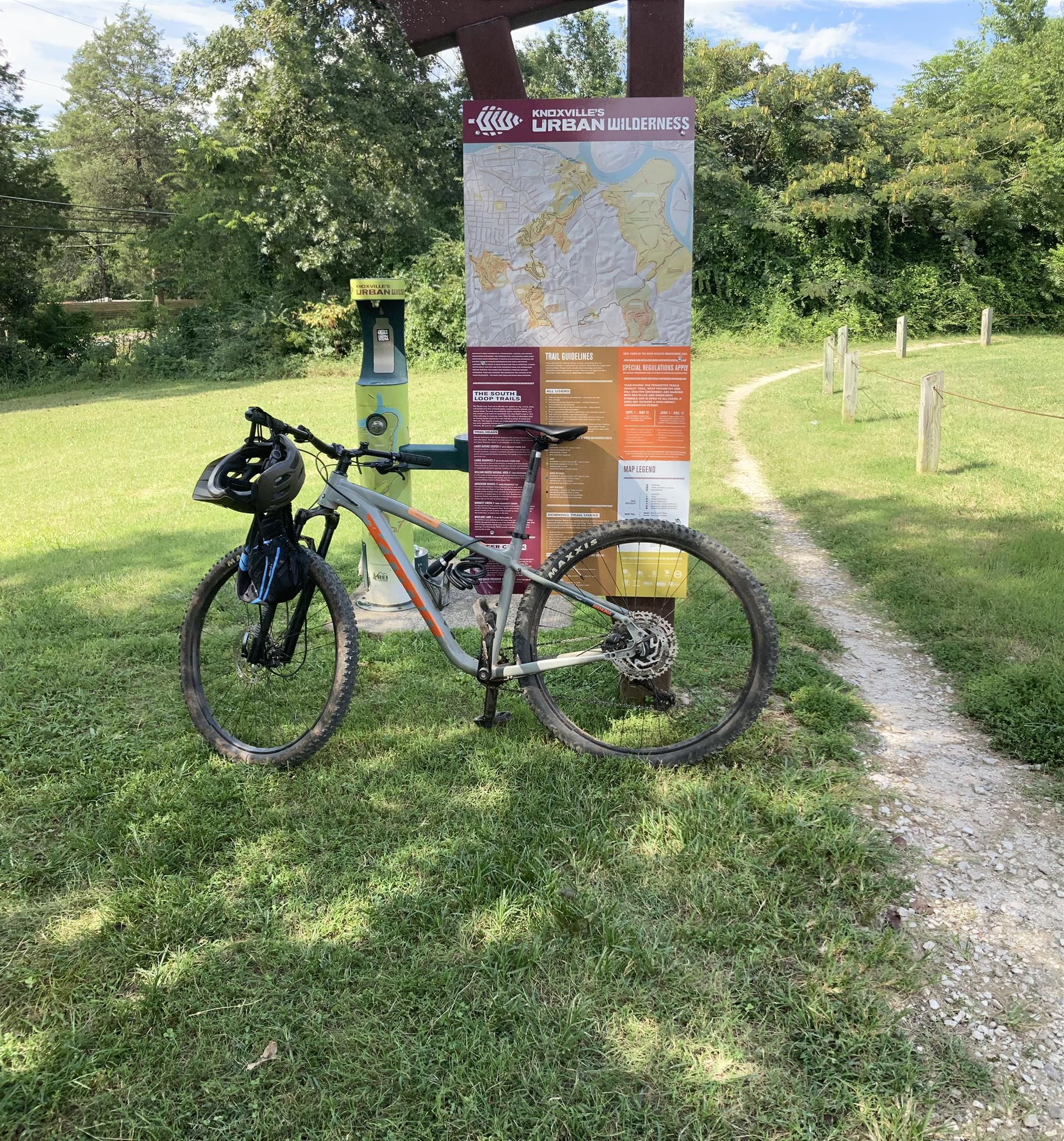 Mountain bike with helmet parked by an information board on a sunny grassy path. - Urban Wilderness loop