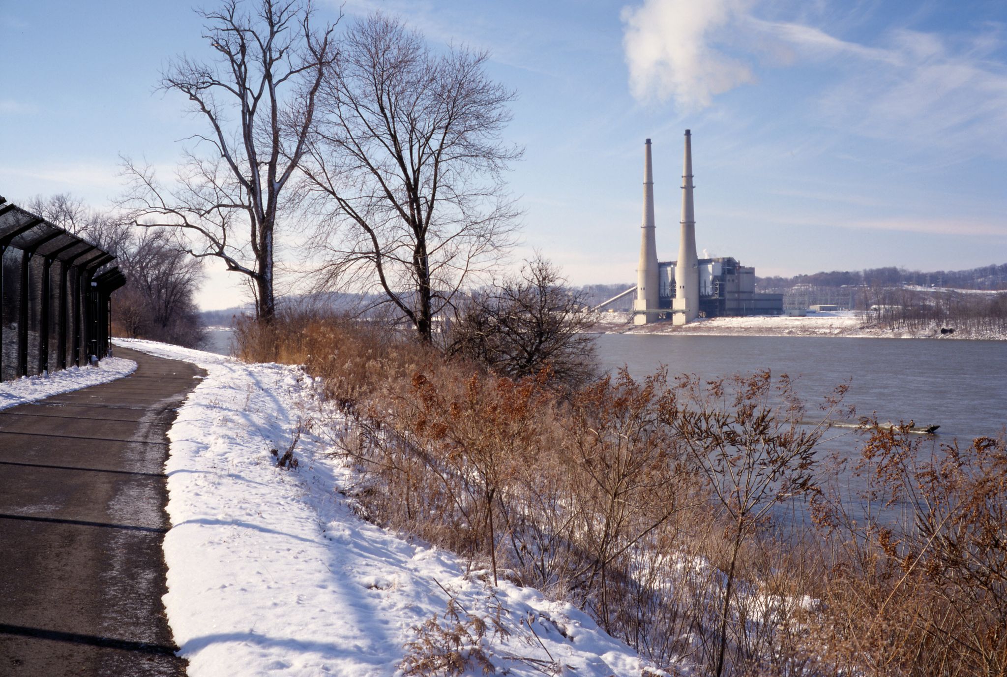 Snowy path next to a river with bare trees and an industrial plant. - Louisville Loop