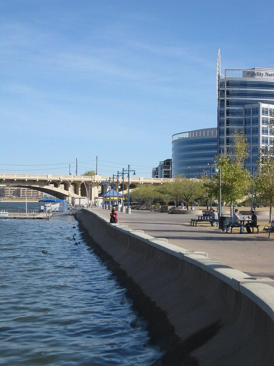 People relax on a sunny waterfront promenade next to a river and buildings.