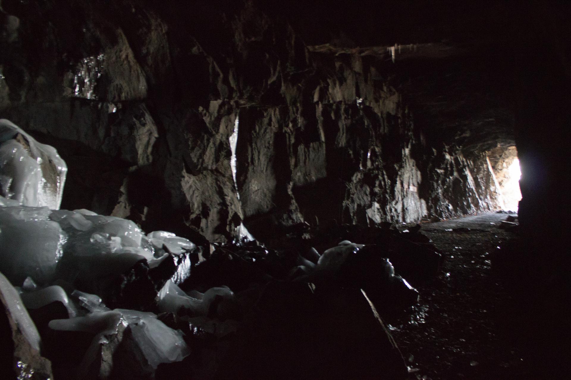 Dark tunnel interior with icy formations on rough rock, light at end.