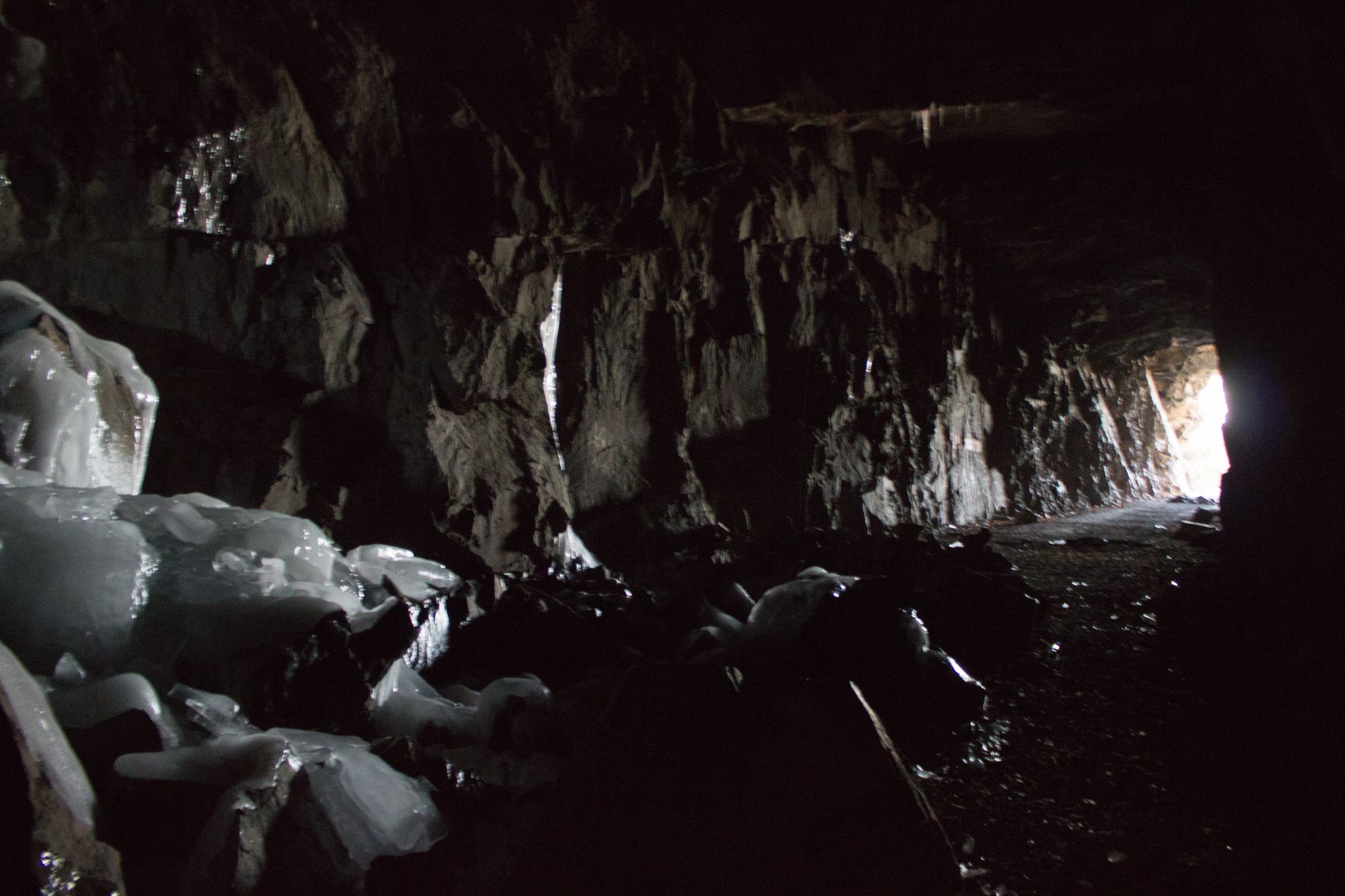 Dark tunnel interior with icy formations on rough rock, light at end. - Loki Trail – Ely's Peak Tunnel loop from Fond du Lac