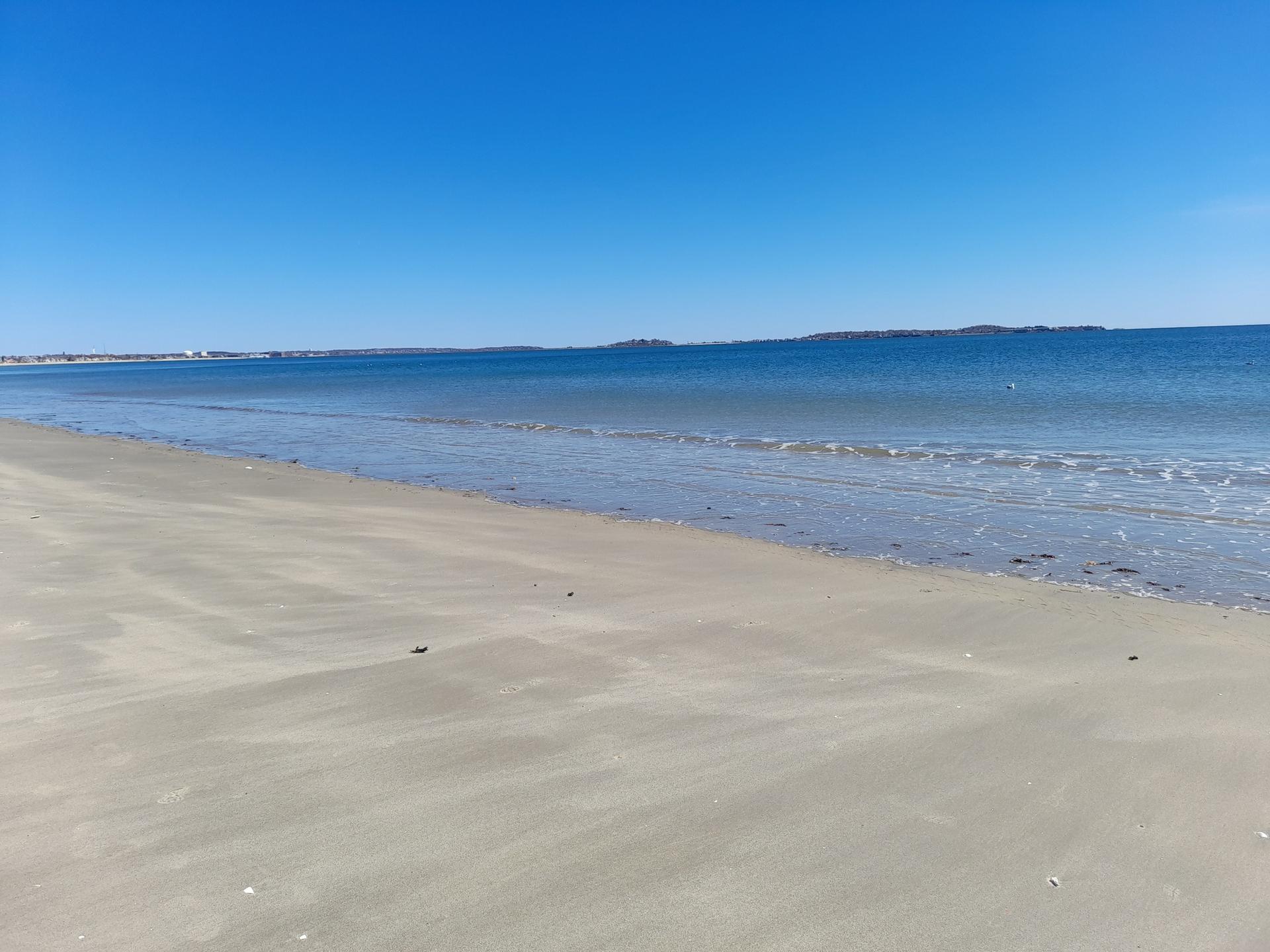 Empty sandy beach with gentle waves and distant land under blue sky.