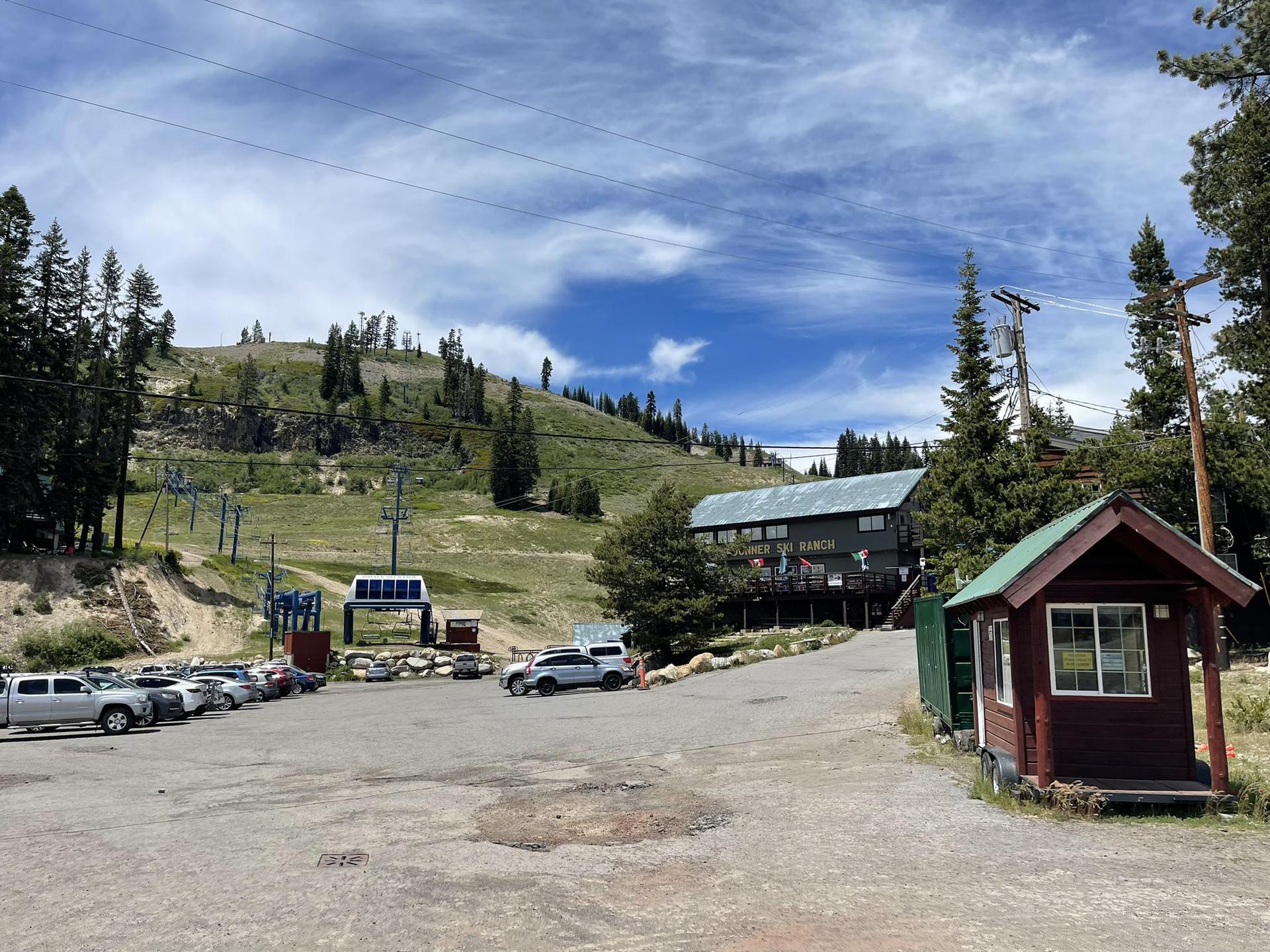 Ski resort base with buildings, parking lot, and grassy slopes under a bright, cloudy sky.
