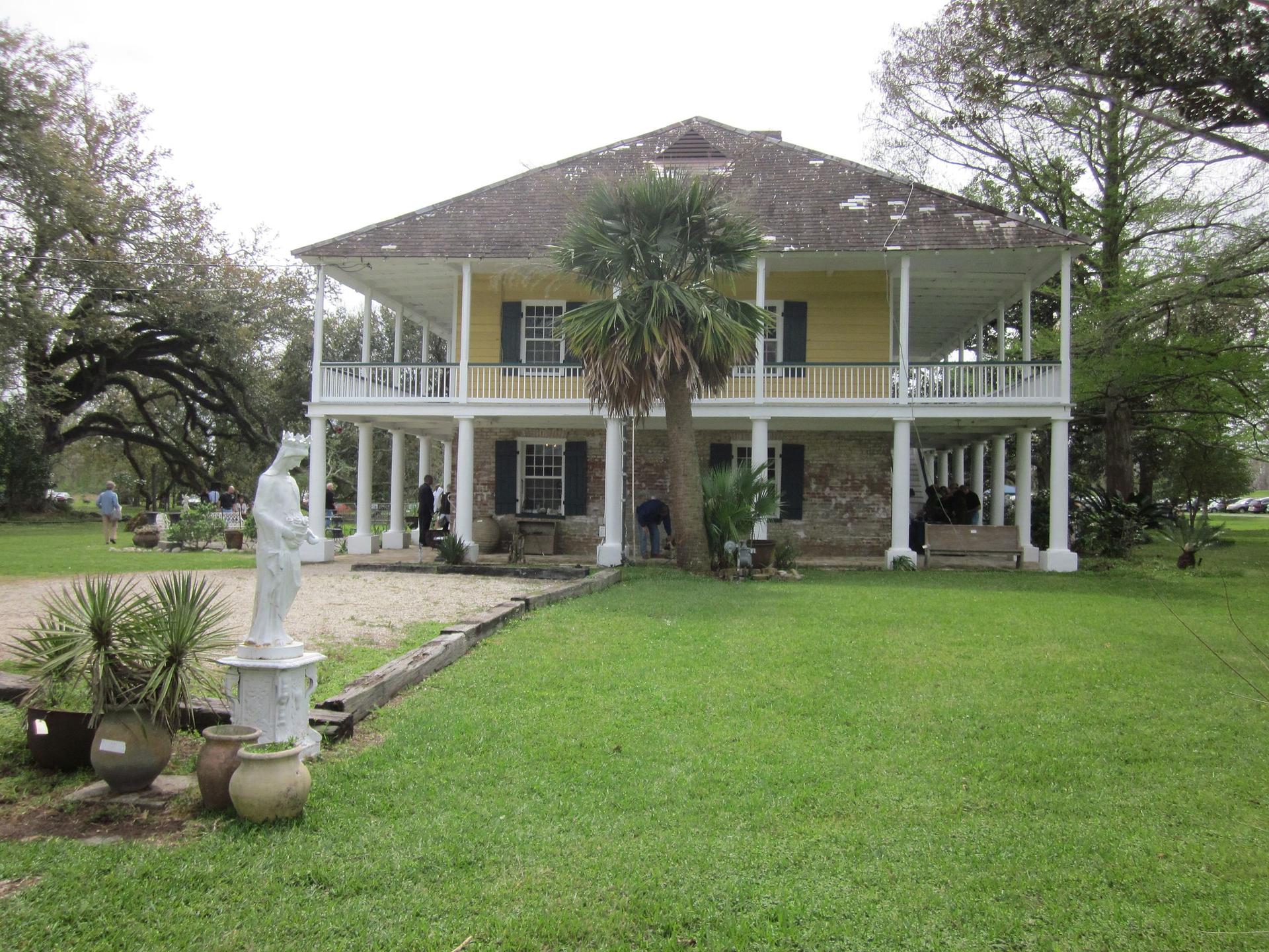 Large yellow house with double porches, palm tree, green lawn, cloudy day.
