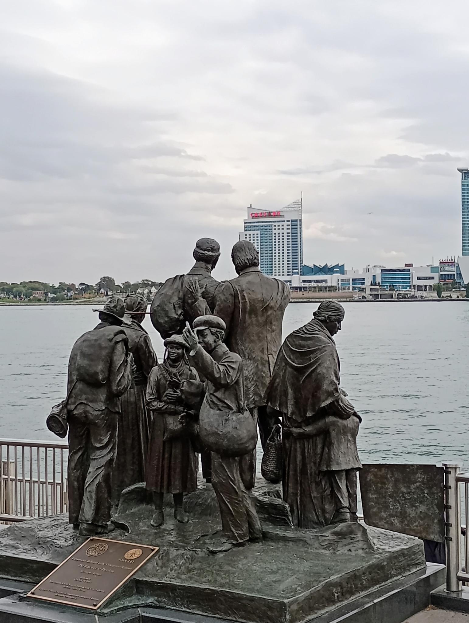 Bronze statue of a group of people by a river, with city in background.
