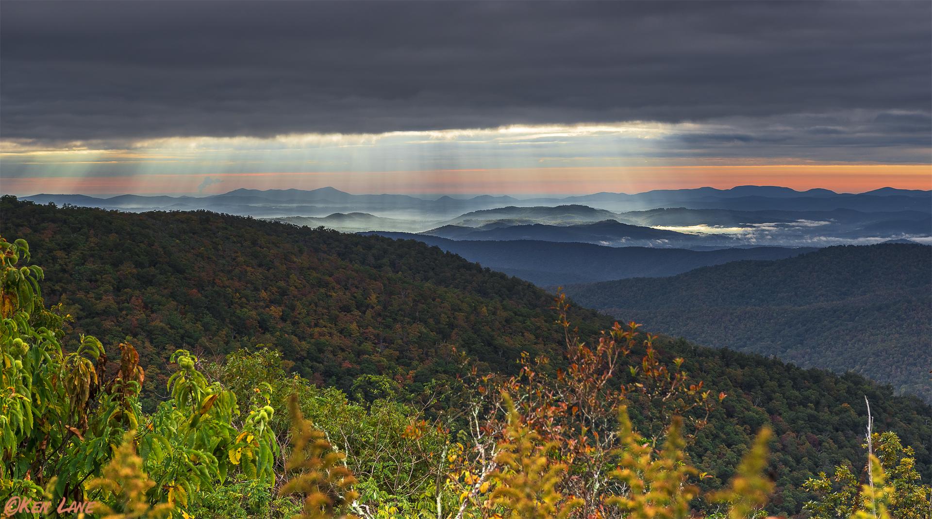 Dramatic sun rays pierce dark clouds over layered mountains at sunrise.
