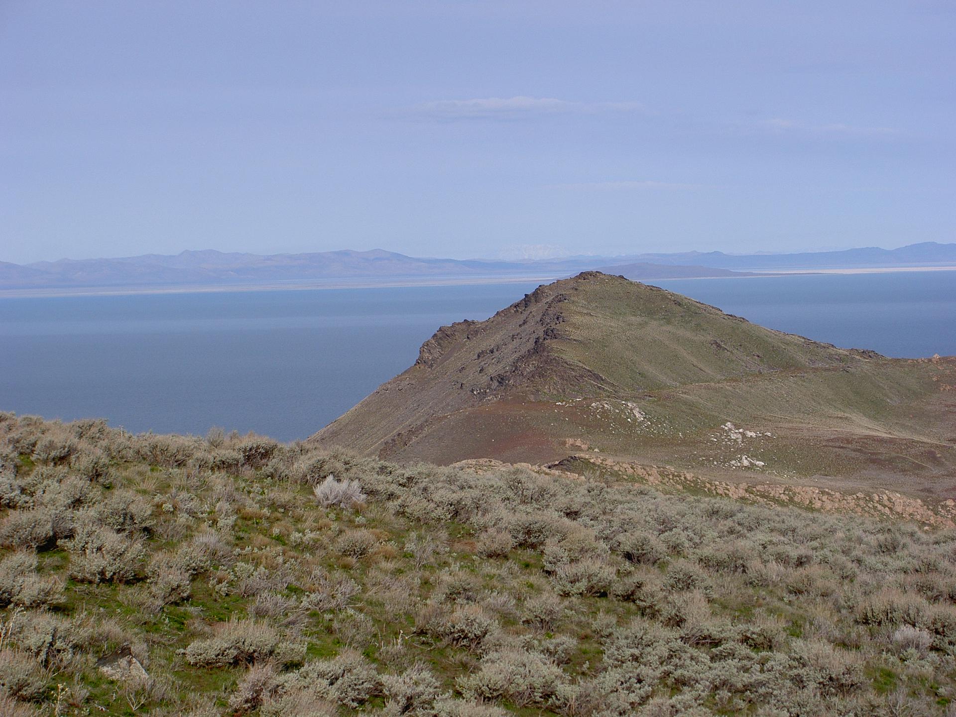Grassy hills overlook a rocky peak and vast, calm blue water.