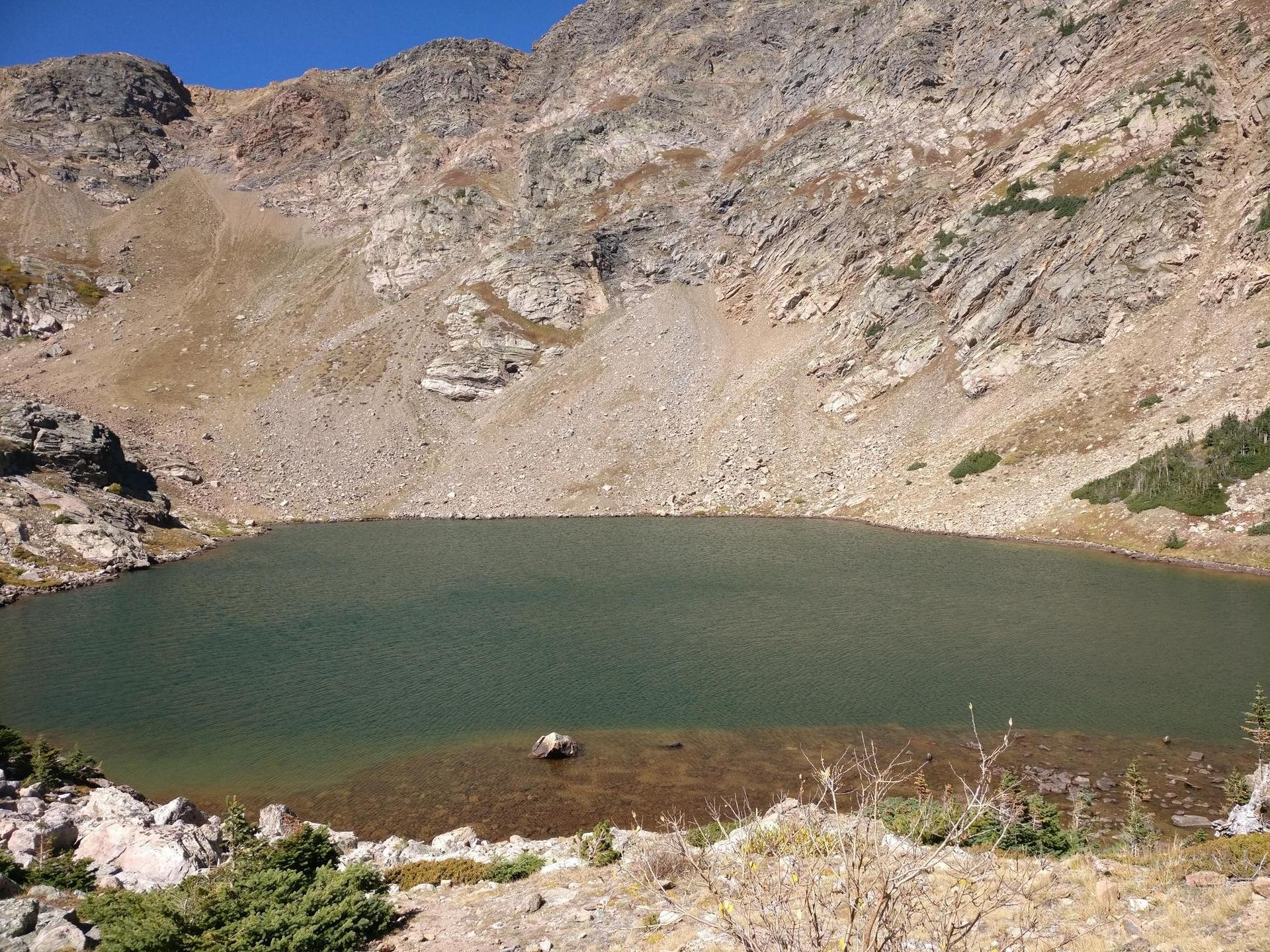 A dark green mountain lake at the base of a steep, rocky, barren mountain.