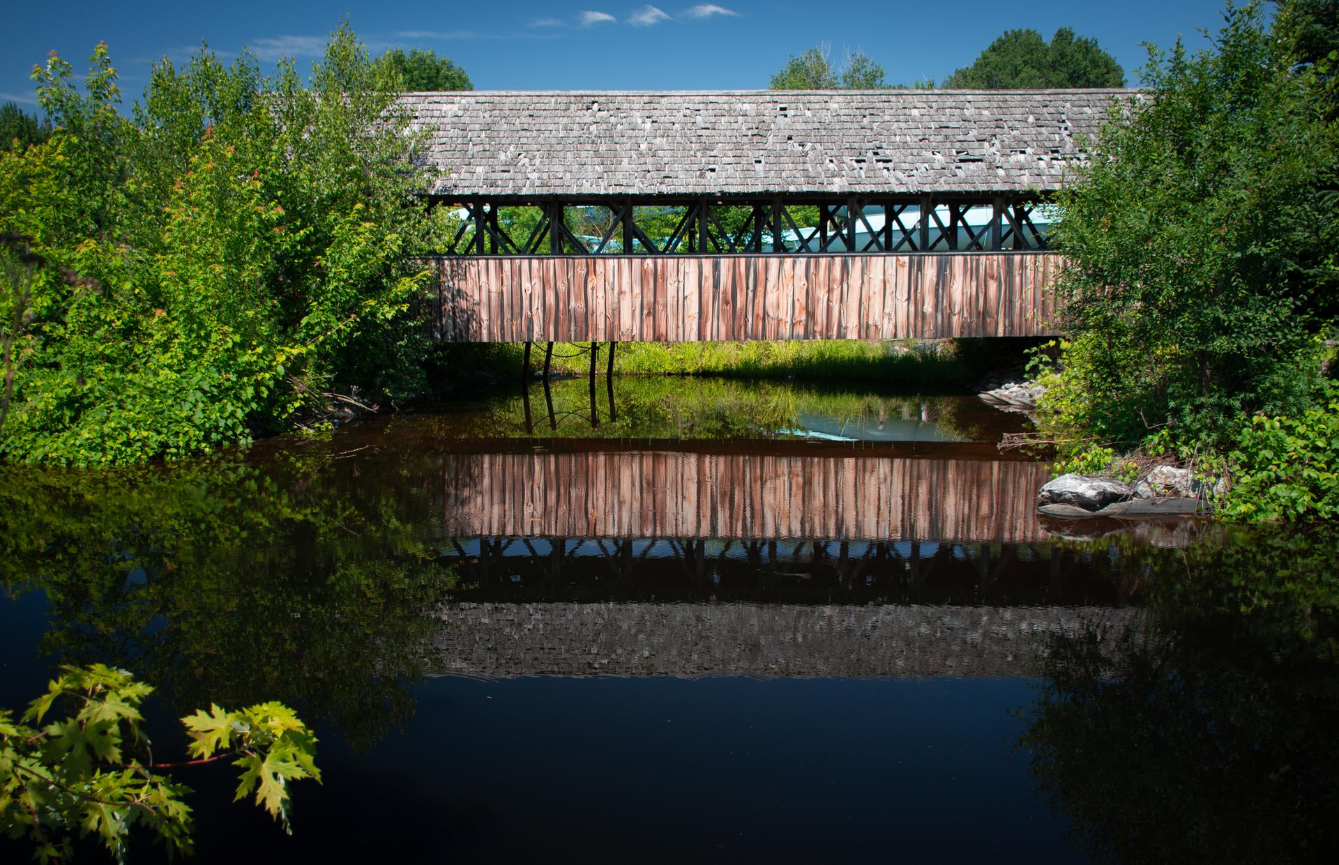 Wooden covered bridge over dark water, surrounded by green trees under a bright sky.