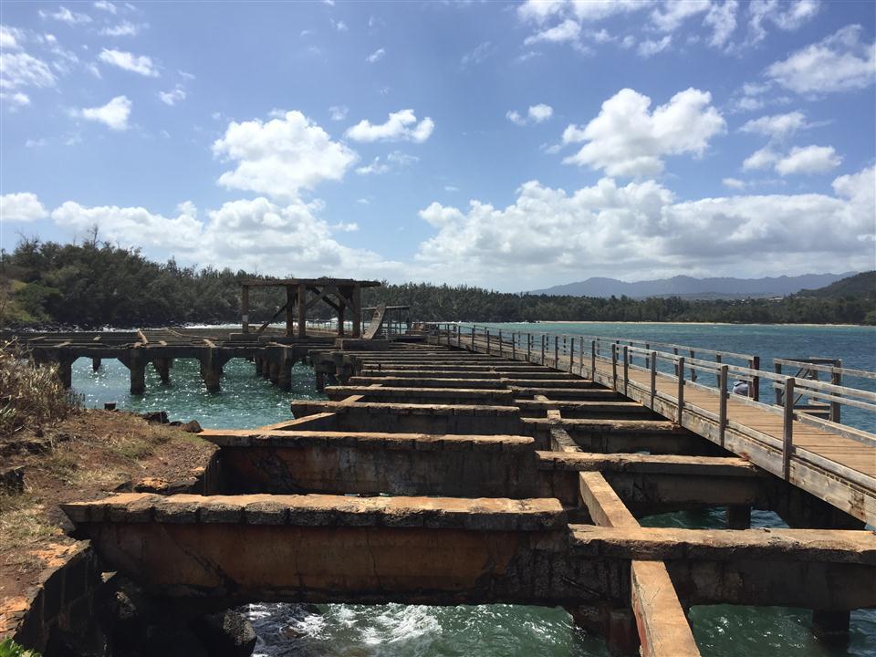 An old pier extends over blue water towards distant mountains on a sunny day.