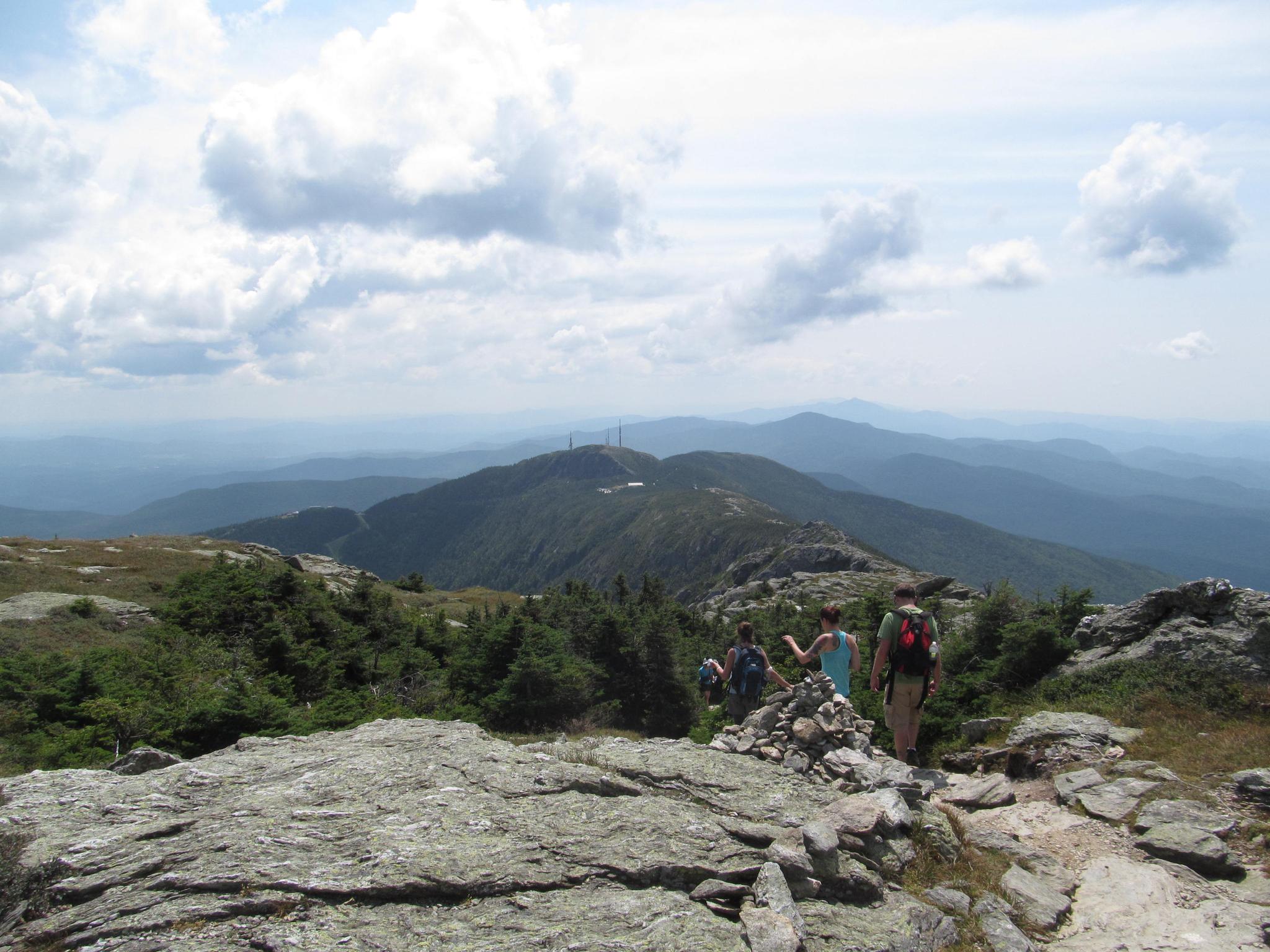 Hikers on a rocky mountain path with distant forested peaks under clouds.