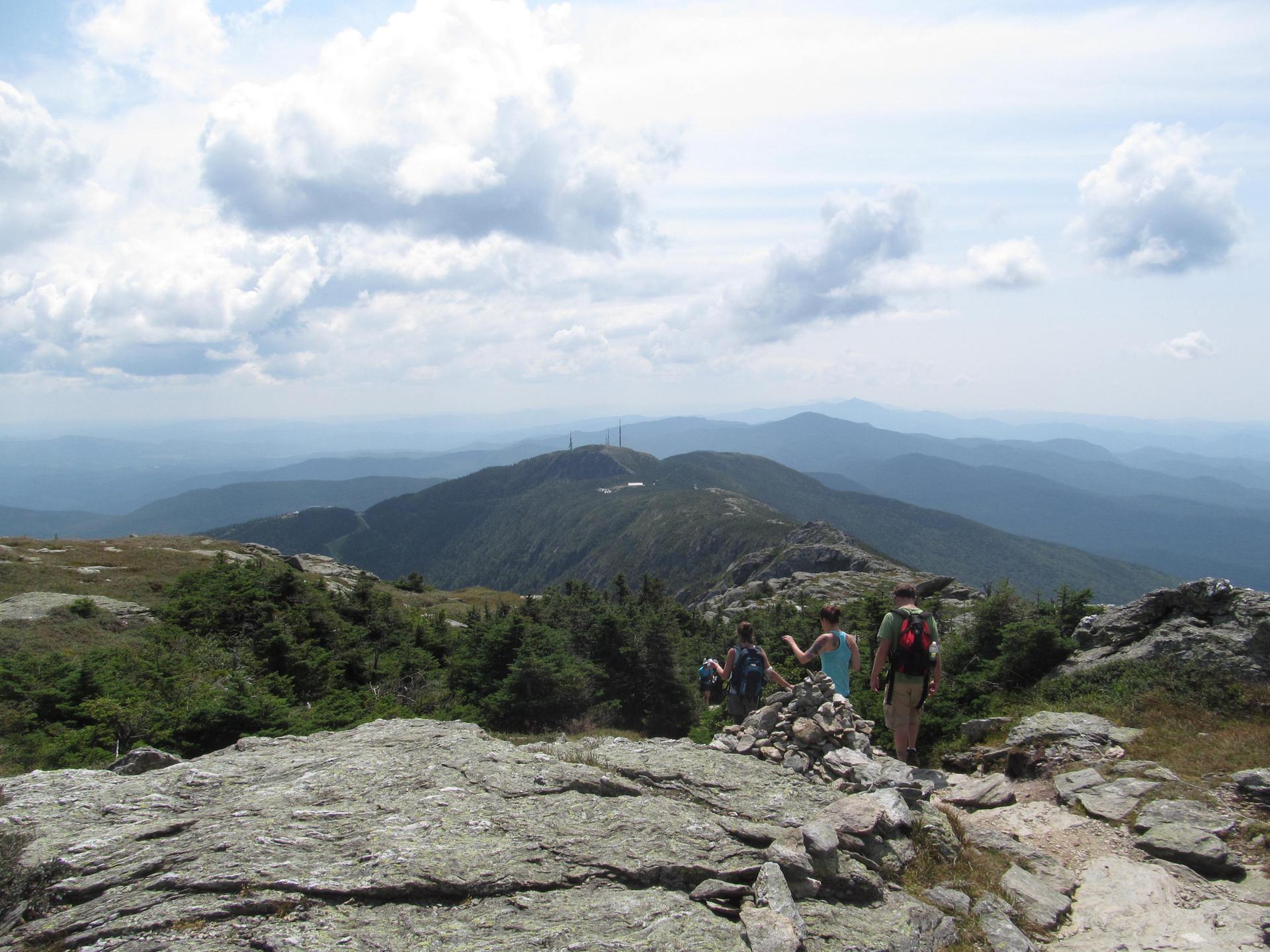 Hikers on a rocky mountain path with distant forested peaks under clouds.
