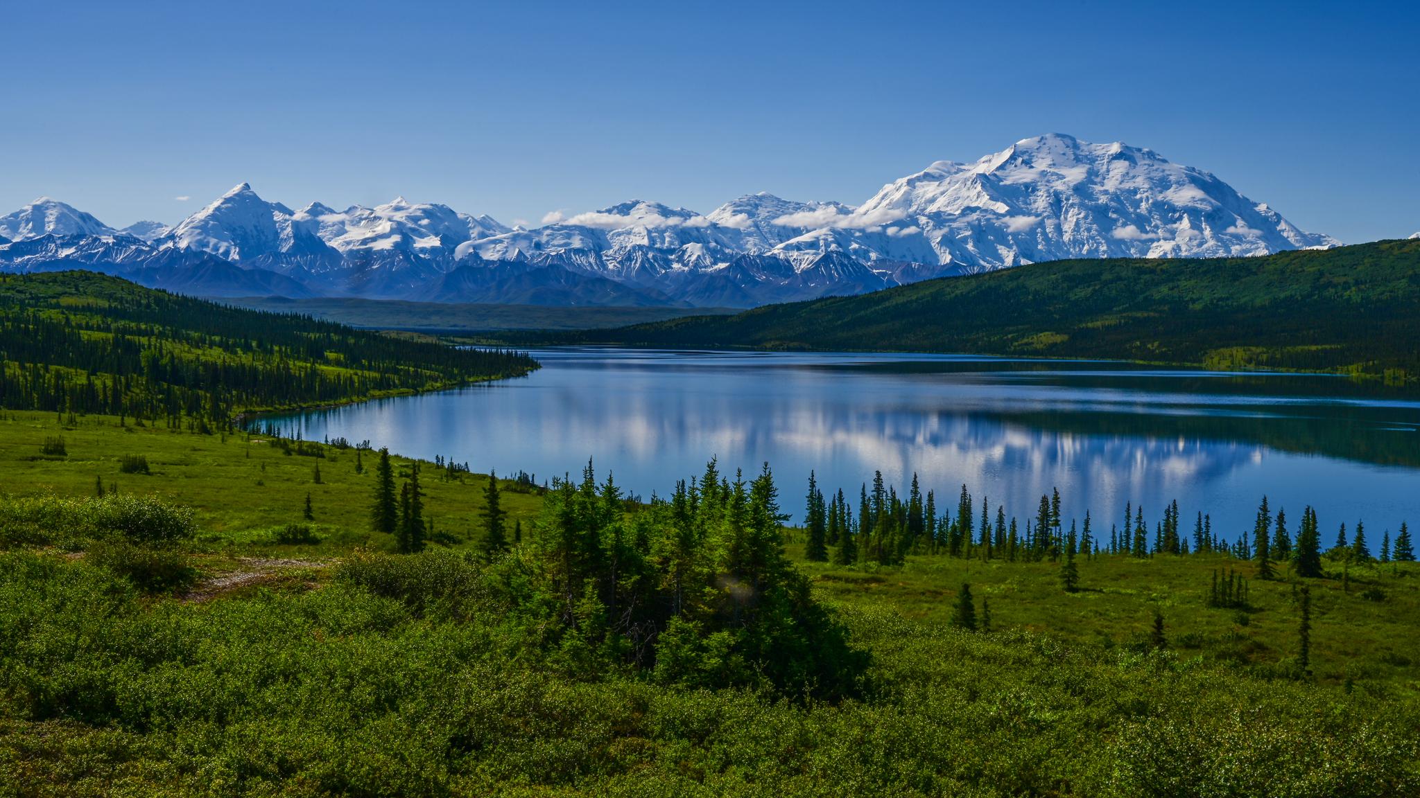 Vast blue lake reflecting snow-capped mountains, surrounded by green forests under clear sky.