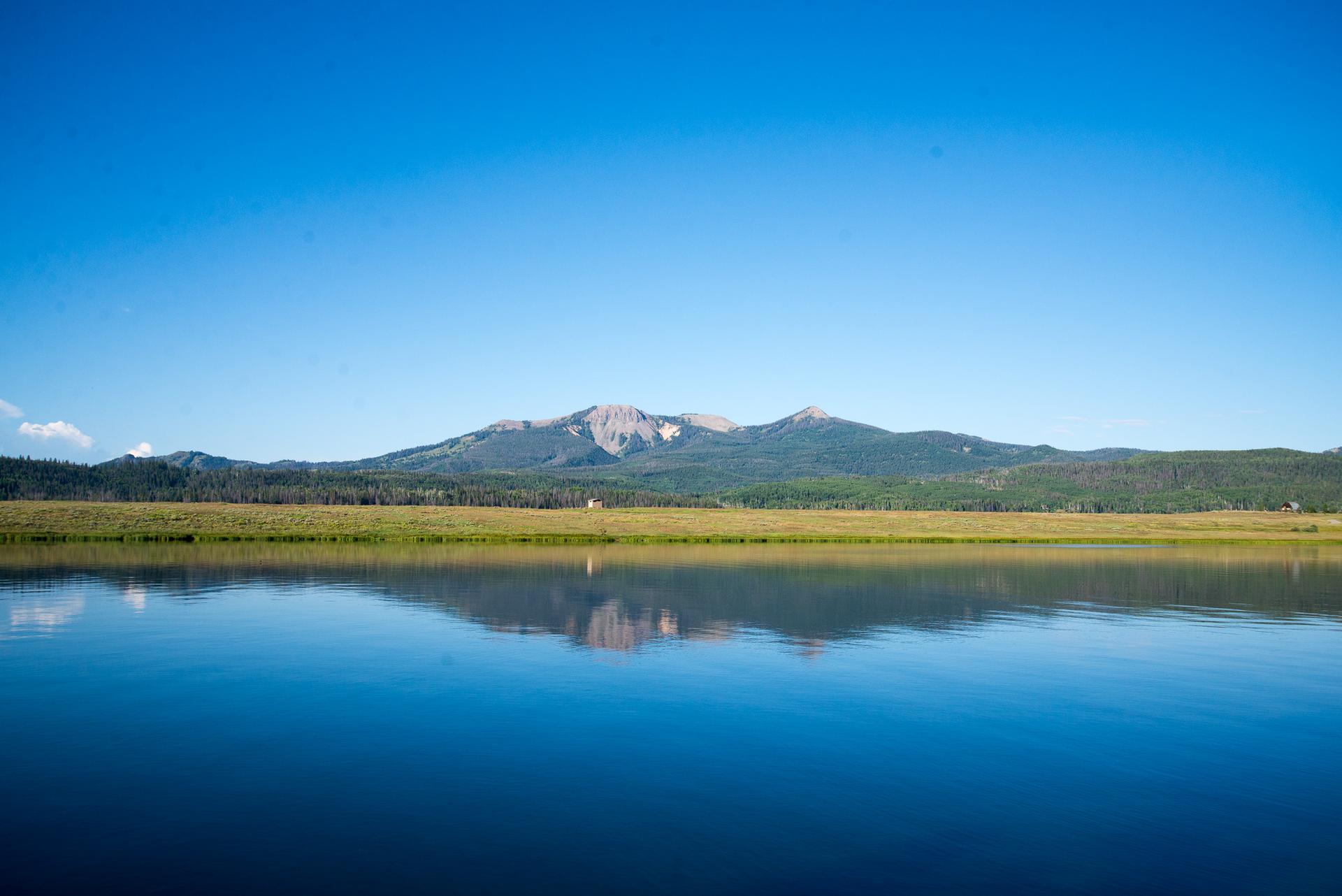 Serene lake reflecting a distant mountain range under a bright blue sky.