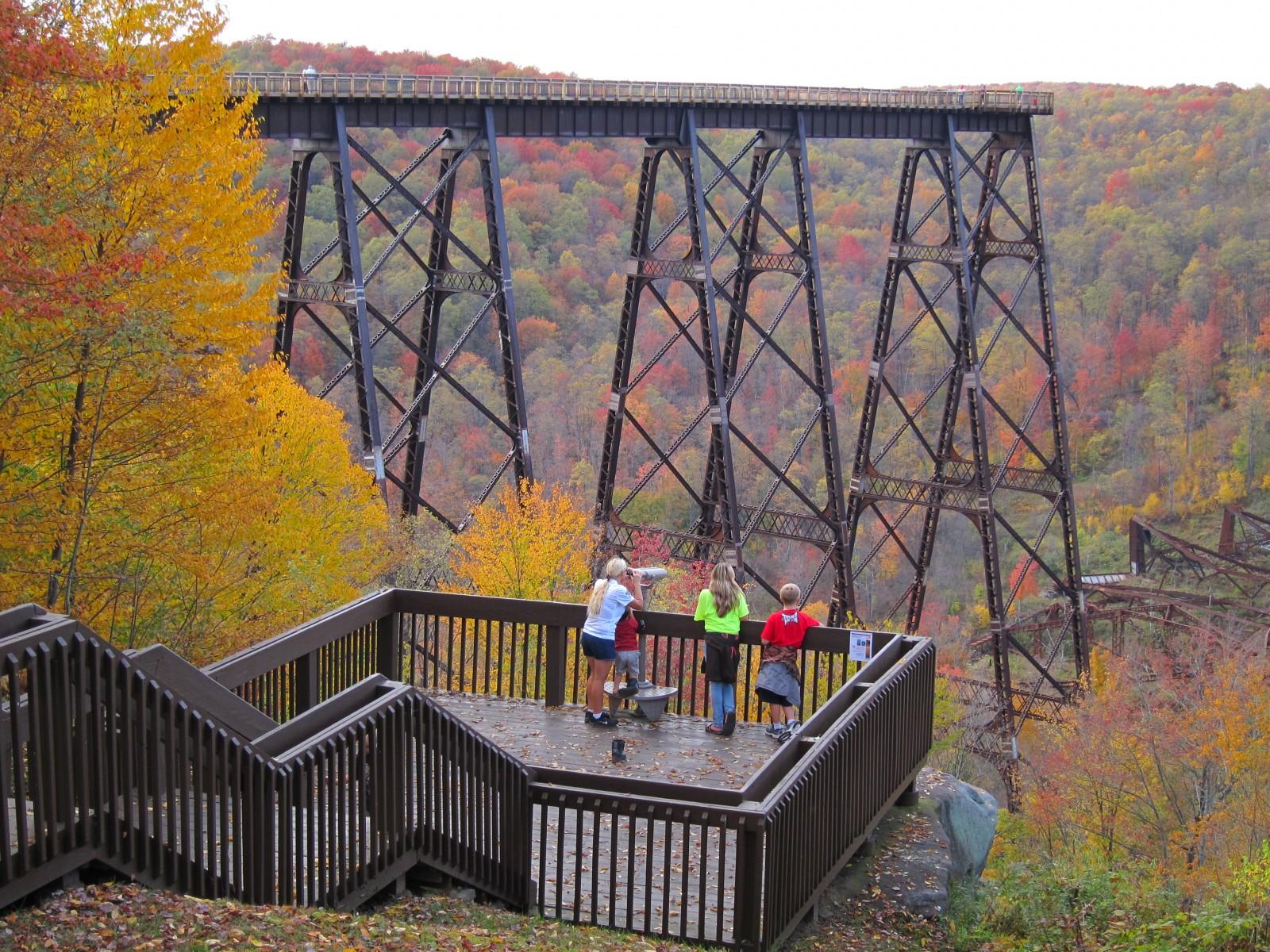 People on a wooden observation deck viewing a massive metal bridge and autumn trees.