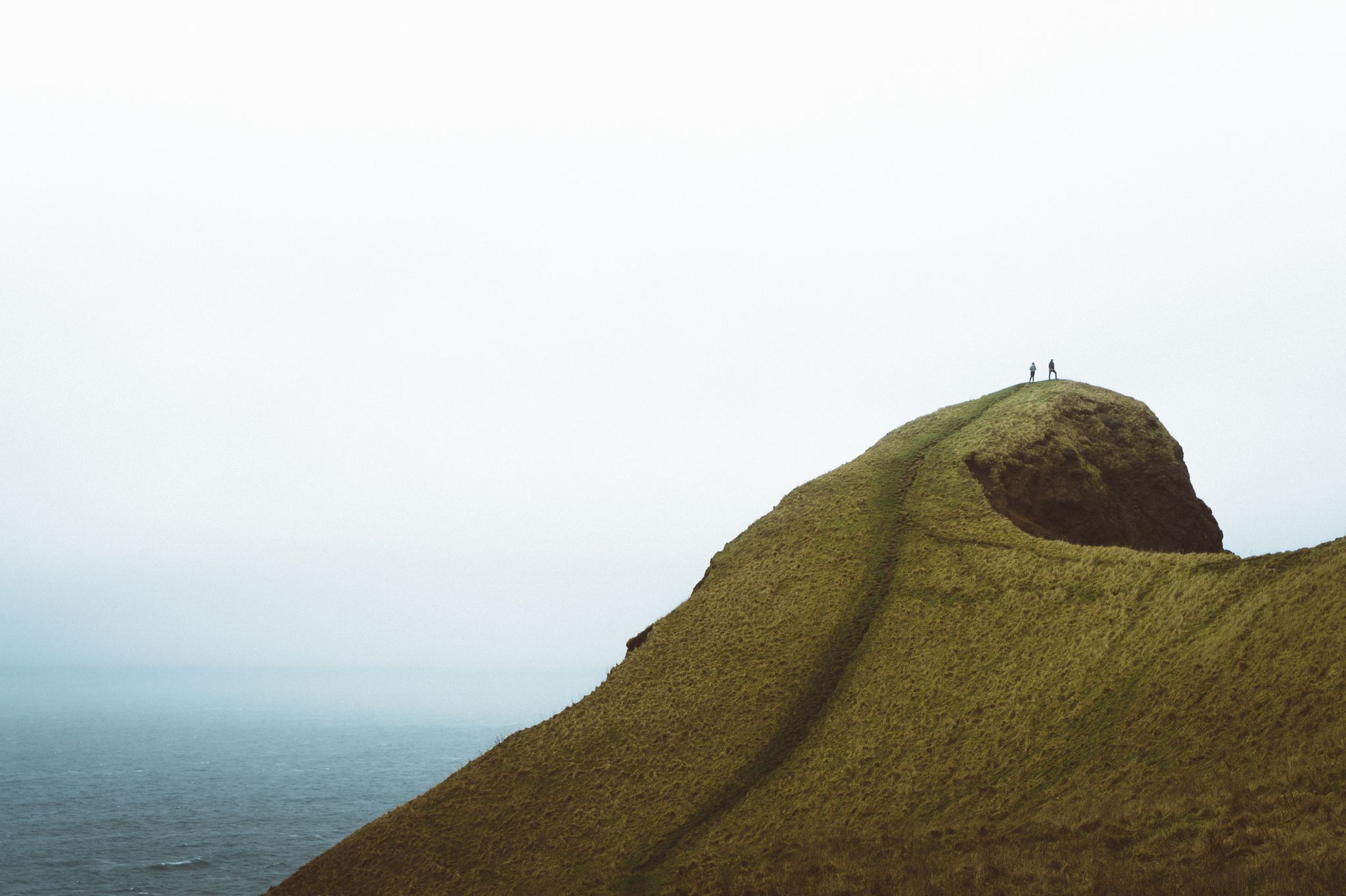 Two small figures stand atop a steep, grassy hill overlooking the ocean. - The Thumb via The Knoll