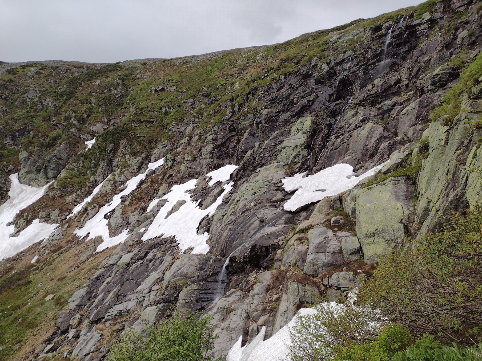 Steep rocky mountain slope with snow patches and small waterfalls under cloudy sky.