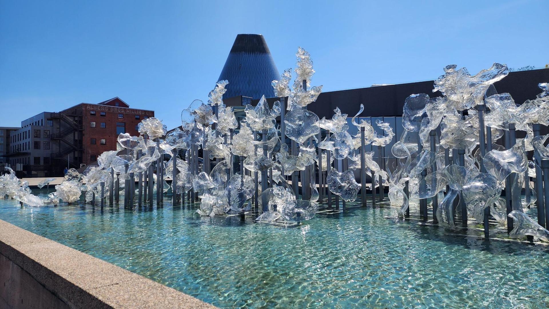 Clear glass sculptures in a water feature under a bright blue sky.