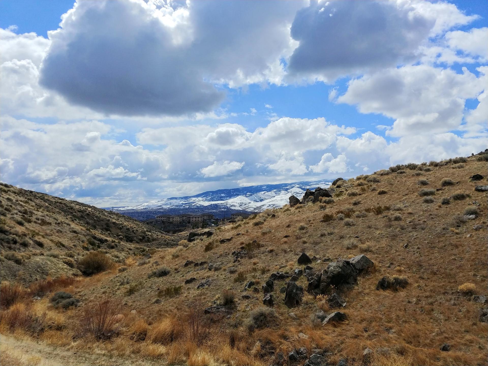 Dry, rocky valley with distant snow-capped mountains under a bright, cloudy sky.
