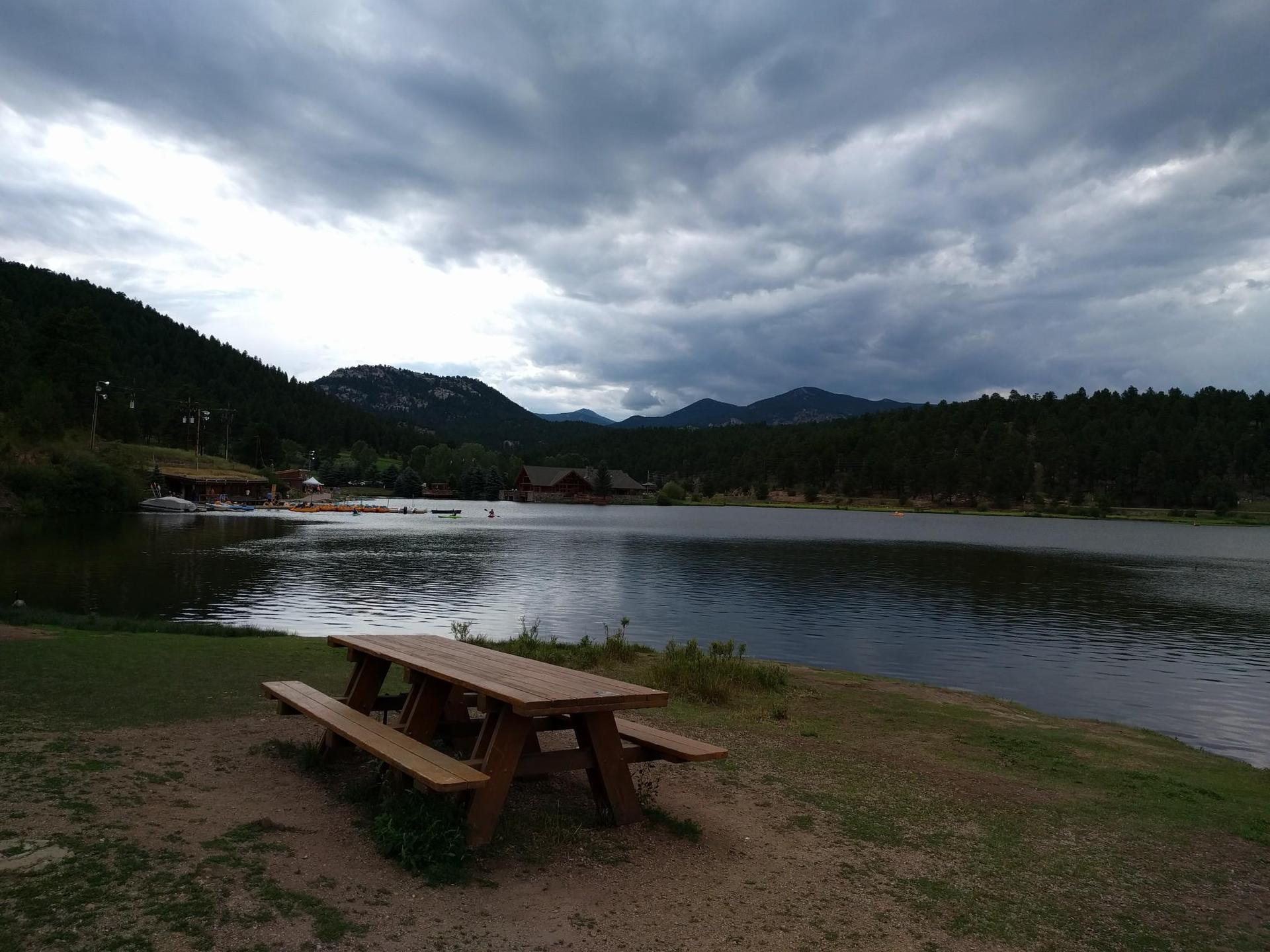 Picnic table by a dark lake with forested mountains under cloudy skies.