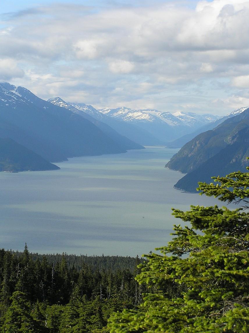 Cloudy day, vast body of water between forested mountains with snowy peaks.