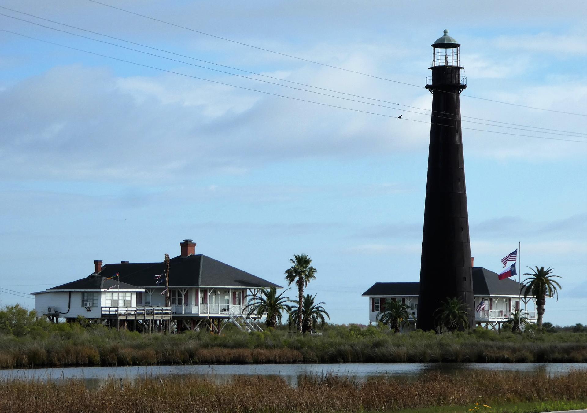 Tall dark lighthouse and white buildings by wetlands under a bright sky.