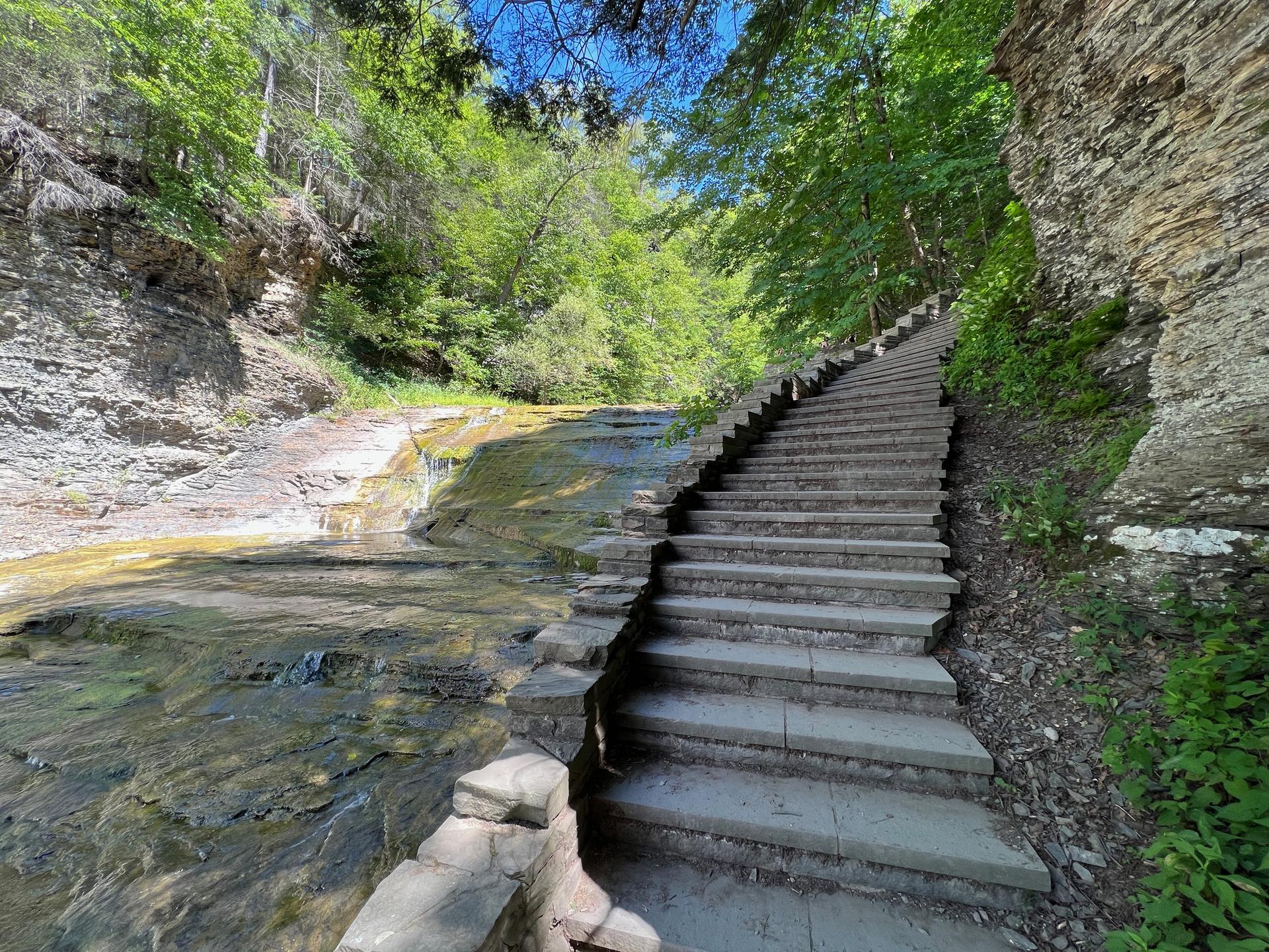 Stone staircase ascends beside a shallow waterfall and rocky cliff in sunlight.